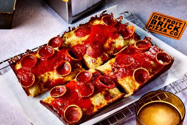 Rectangular pepperoni pizza with melted cheese and tomato sauce, served on a wire rack with a side of dipping sauce and a Detroit Brick Pizza Co. label in the background.
