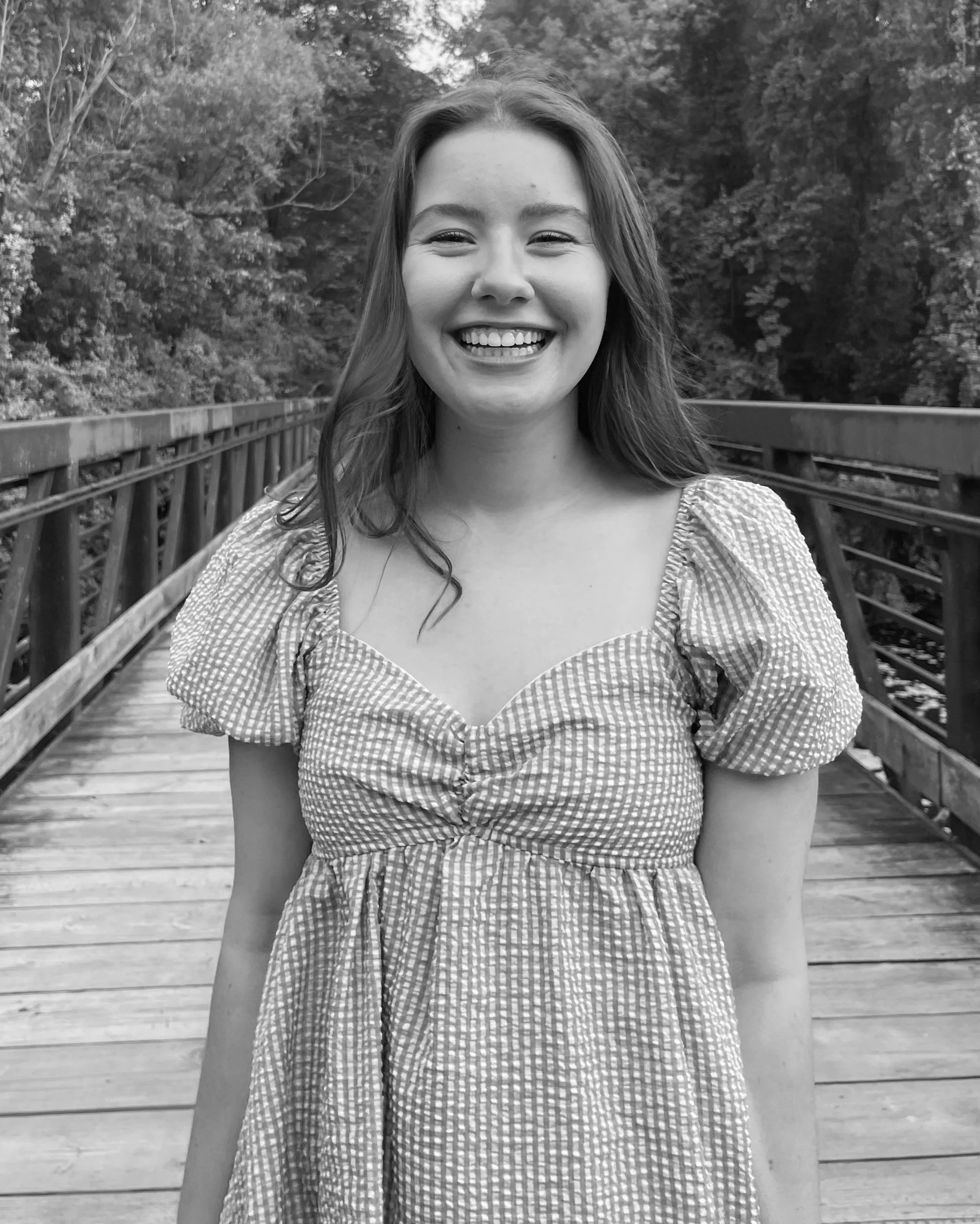 A smiling woman with long hair standing on a wooden bridge with a railing, surrounded by trees in the background.