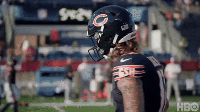 An American football player with long hair, wearing a black helmet and jersey with red, white, and blue details, standing on the field during a game.