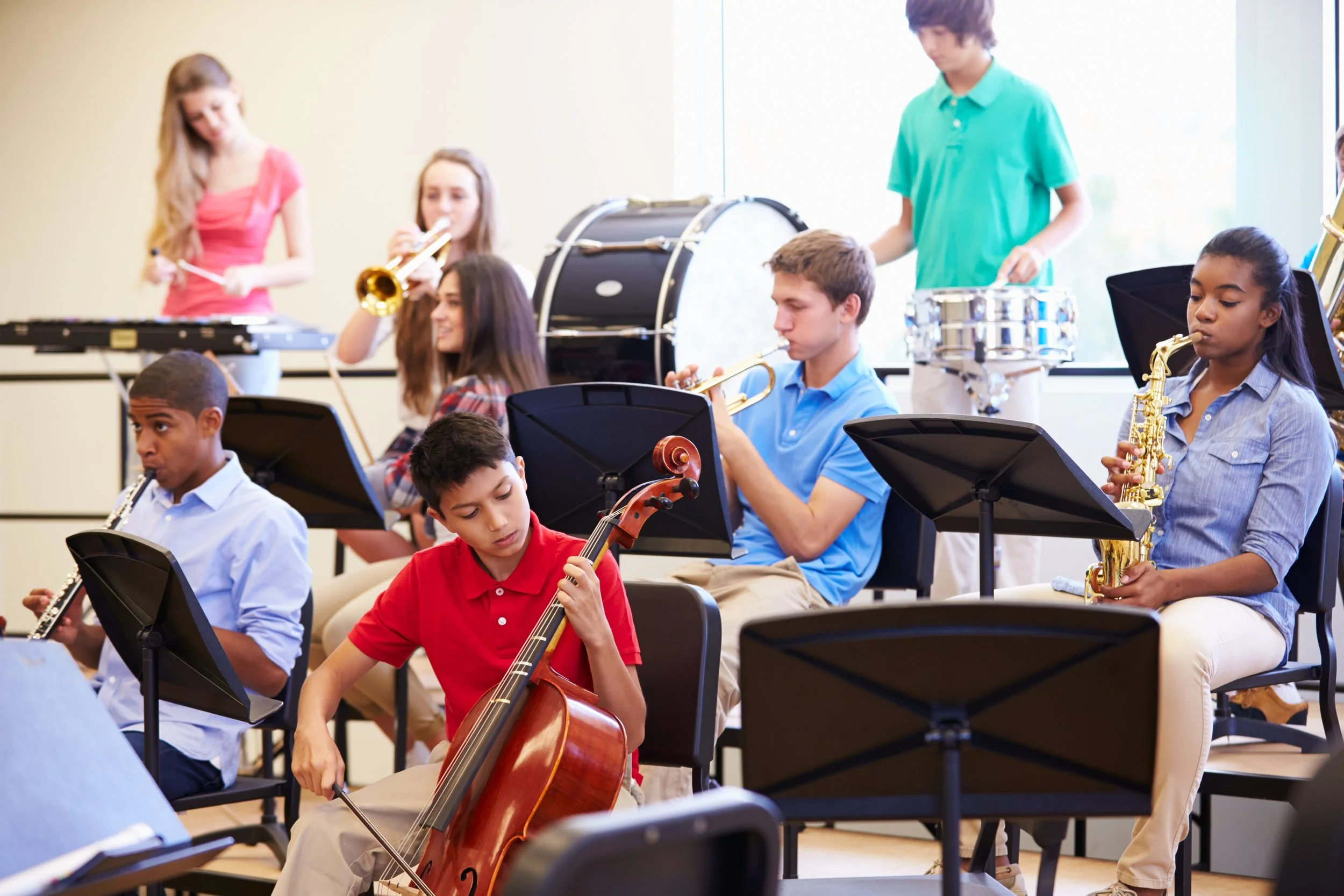 A diverse group of students playing instruments in a school band class, including trumpet, cello, saxophone, oboe, and percussion instruments.