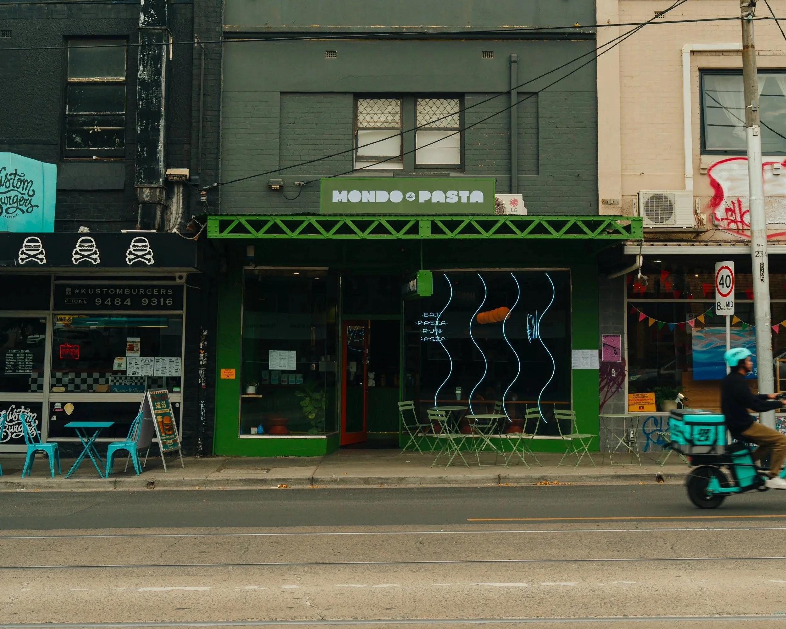 storefront with a green sign reading 'Mondo Pasta,' outdoor seating with green chairs, and a black neon sign inside the window with stylized steam lines at mondo di pasta in melbourne.