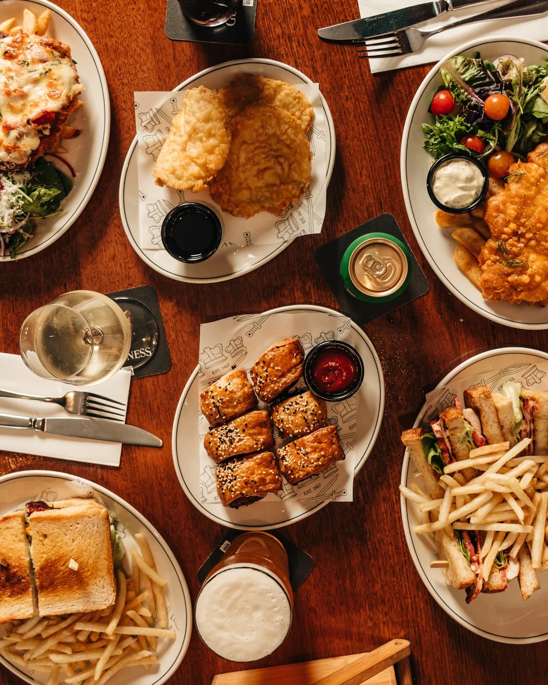 A variety of dishes on a wooden table, including salad, fried chicken, sushi, sandwiches, and drinks.