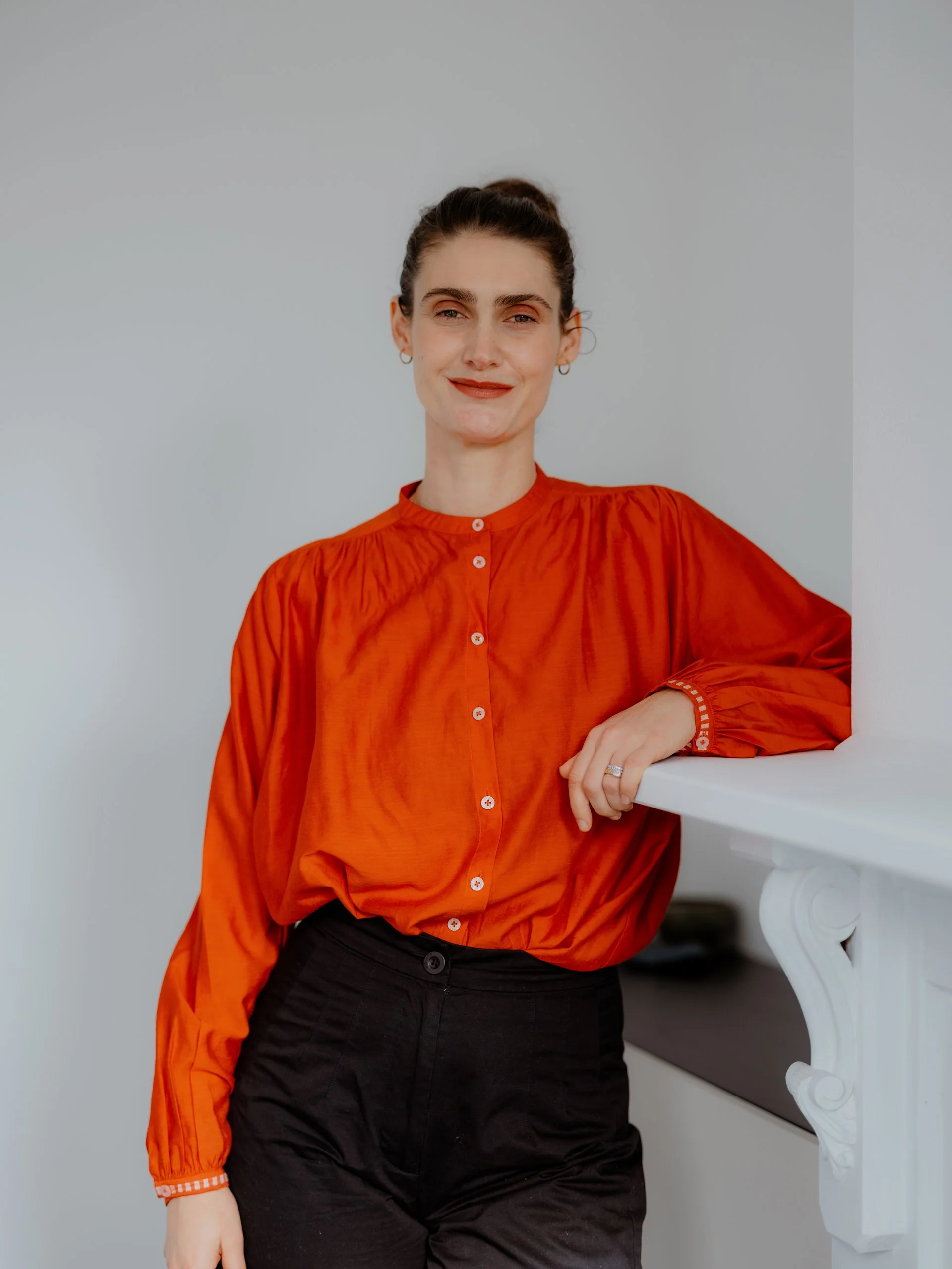 Practitioner, Jo Sharkey, with dark hair in a bun, wearing a red ruffled blouse and black pants, leaning on a white ledge against a plain gray background.