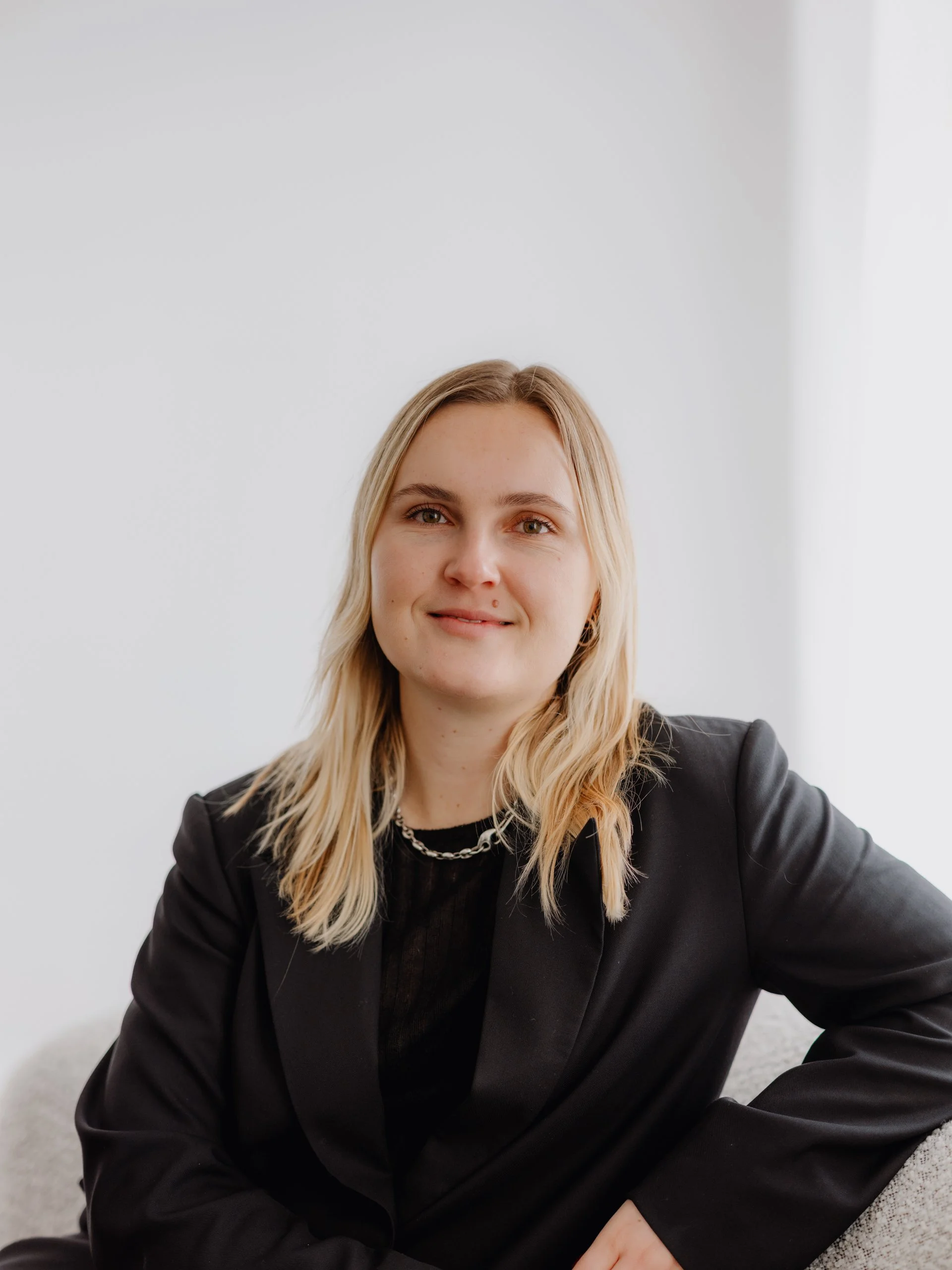 Practitioner, Meghan Smith, with blonde hair, wearing a black blazer and a chain necklace, sitting against a plain white background.