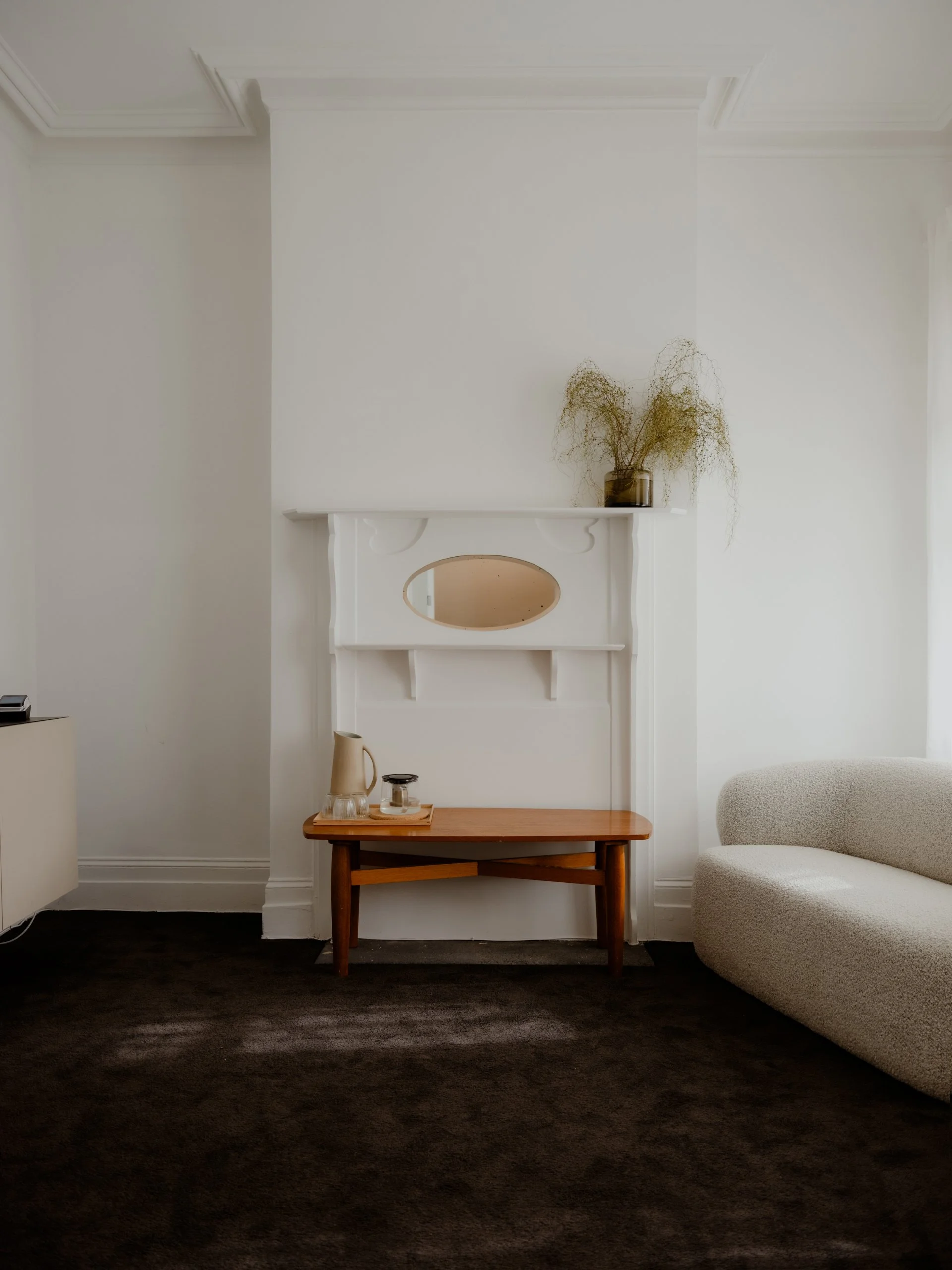 Minimalist living room with a white fireplace mantel, a wooden coffee table, a beige upholstered armchair, and a brown carpeted floor. A clear glass vase with dry, curly plants sits on the mantel.