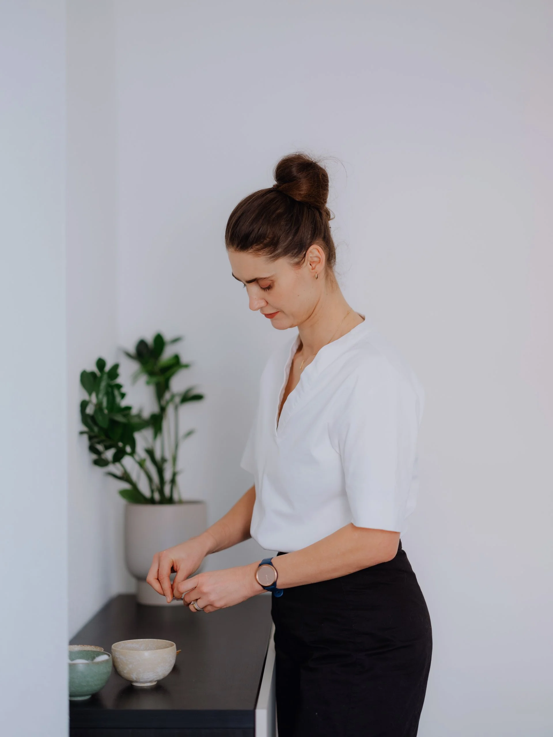 A woman with brown hair in a bun wearing a white shirt and black skirt stands by a dark table with ceramic bowls in front of a light-colored wall with a small green plant in a white pot.