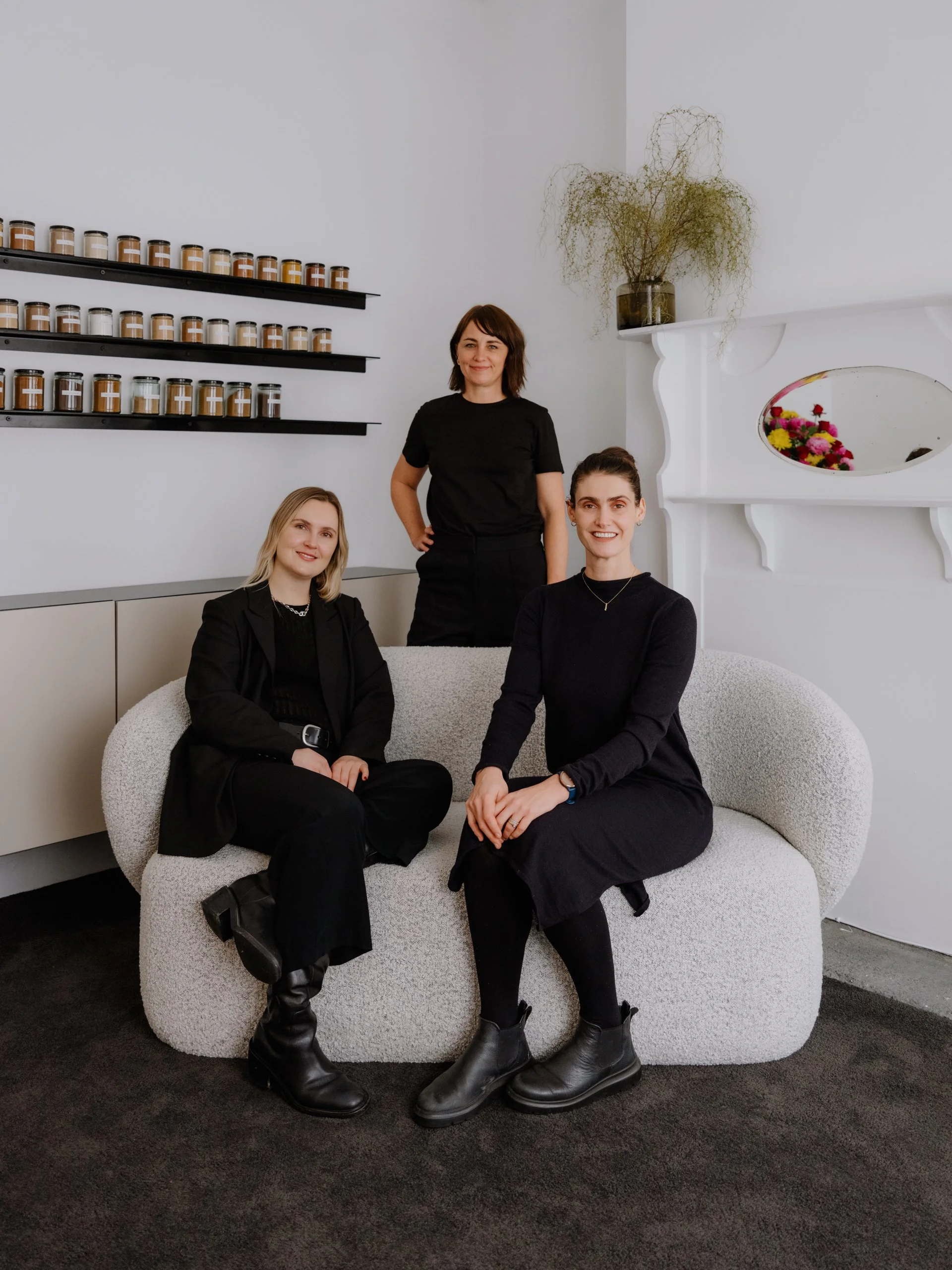 Three women in black clothing sitting and standing on a curved white sofa in a bright room with white walls. Meg & Jo are seated on the sofa, and Kim stands behind them. There are shelves with Chinese herbs and a vase with dried plants on the wall.