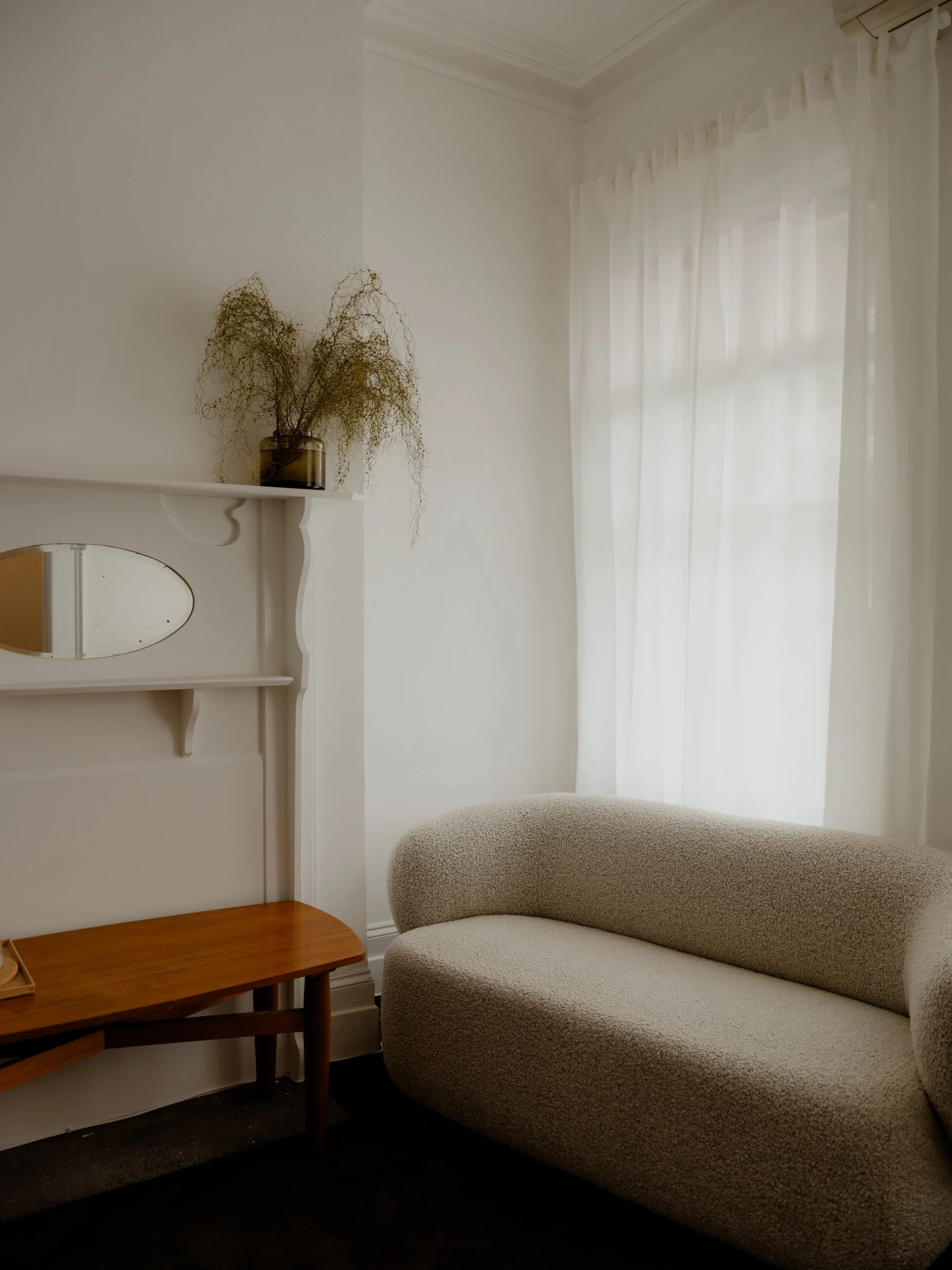 A cozy corner of a room with a beige, curved sofa, a small wooden coffee table, a white shelf with a vase of dried plants, and sheer white curtains letting in natural light.