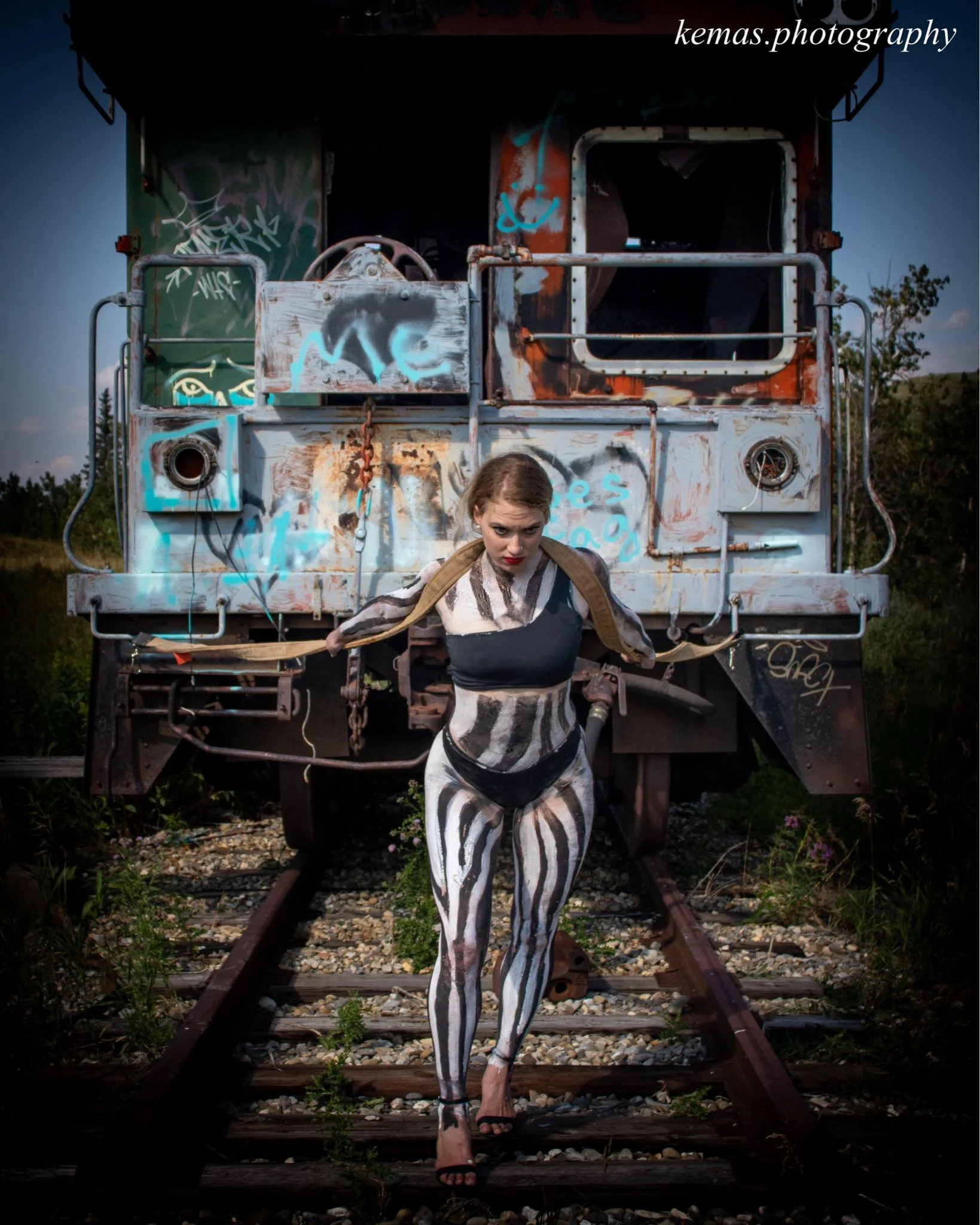 Woman with striped body paint standing in front of a graffiti-covered train caboose on railroad tracks.