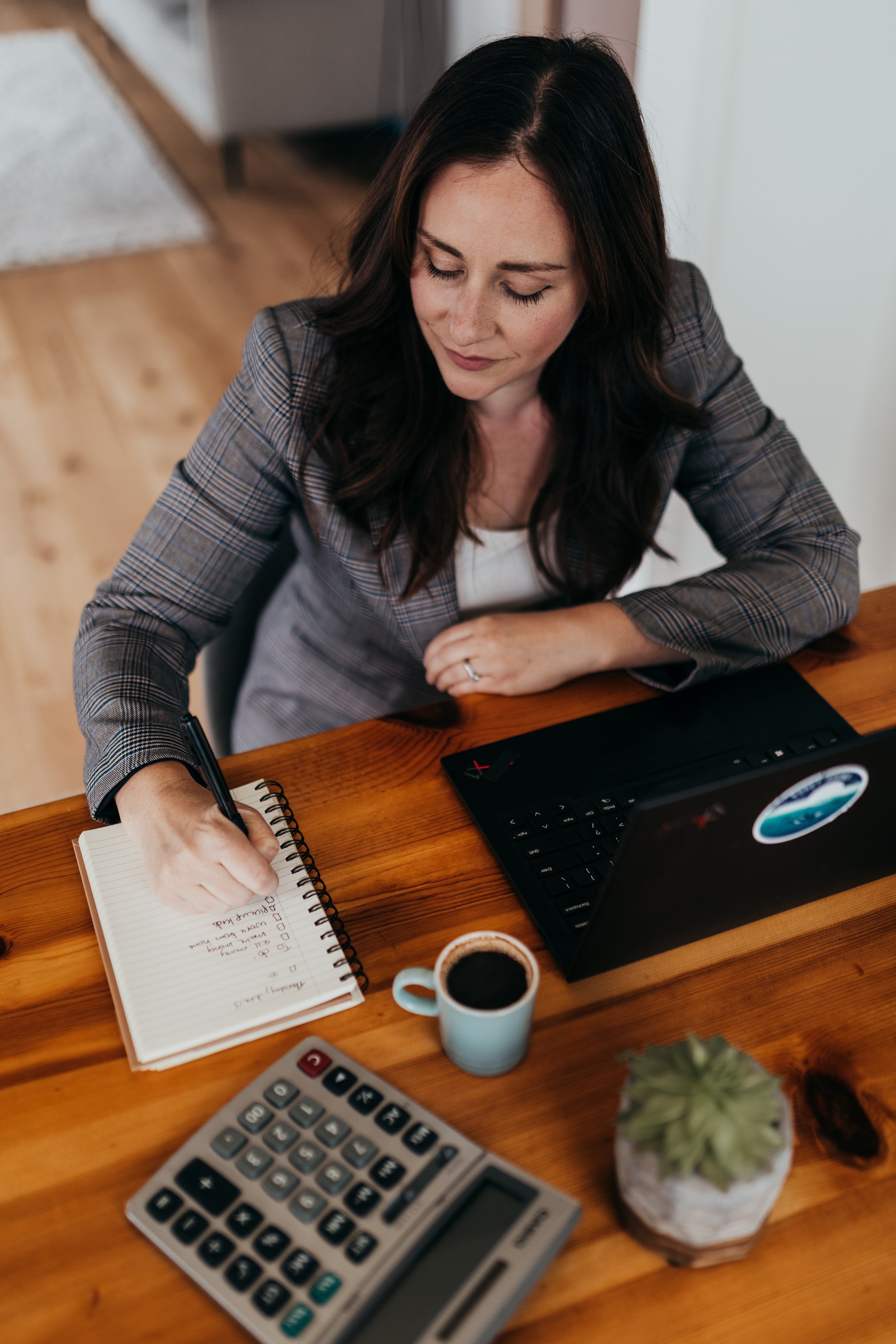 Woman working at a desk with a laptop, notebook, coffee, calculator, and potted plant