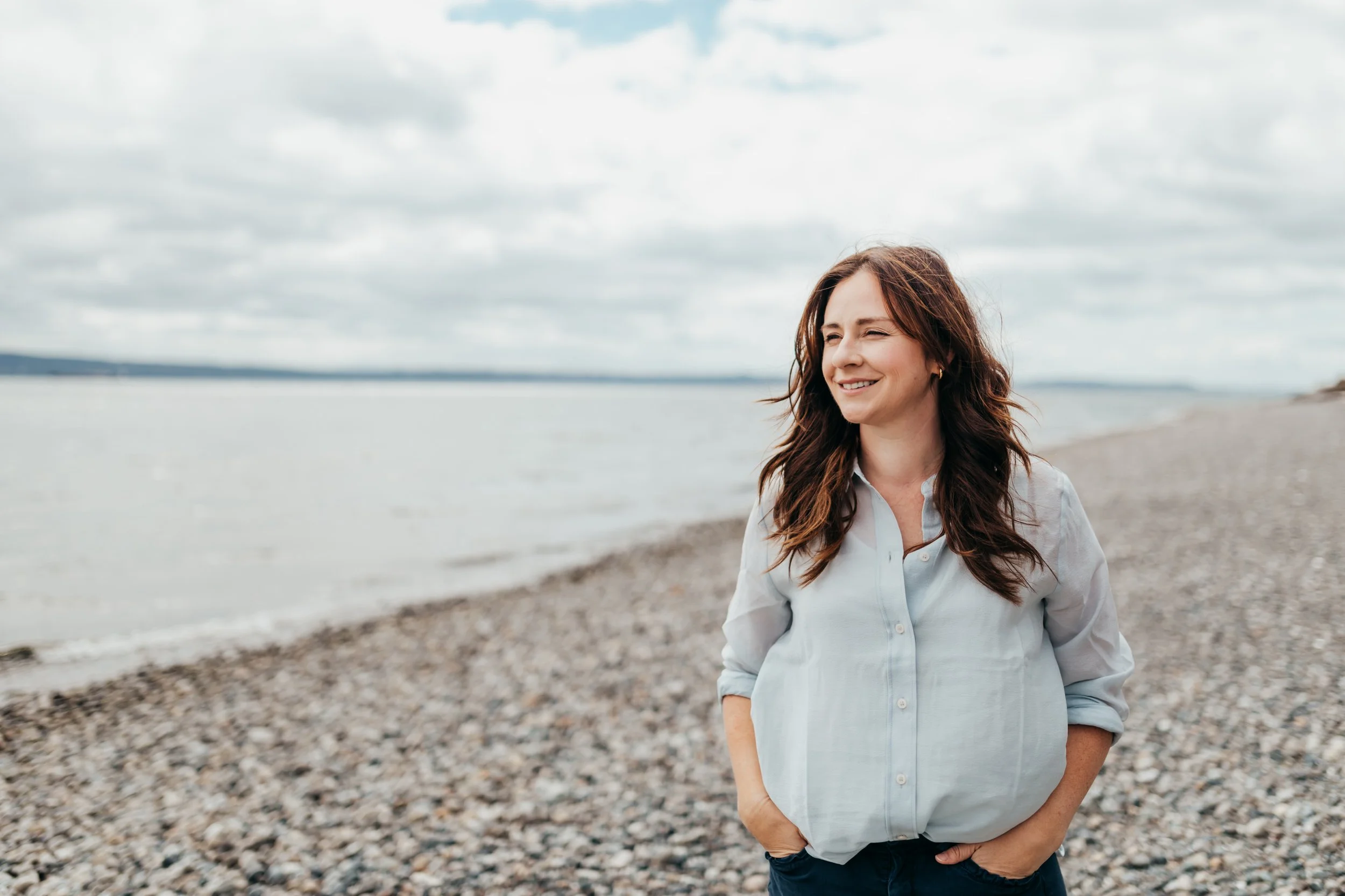 Smiling woman in a light blue shirt standing on a rocky beach beside calm water under a cloudy sky