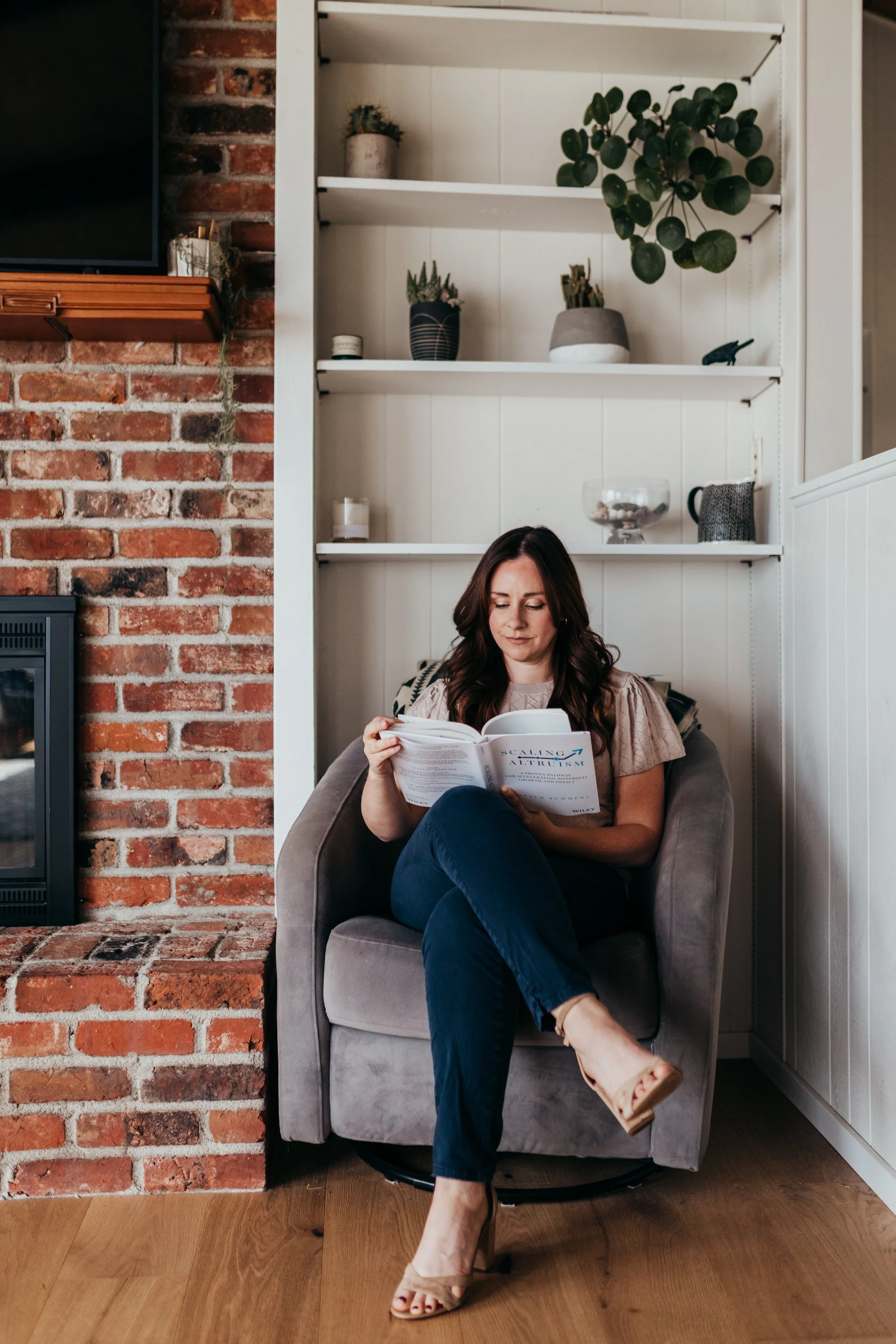 Woman sitting in an armchair reading a book beside a brick fireplace and built-in shelves with houseplants.
