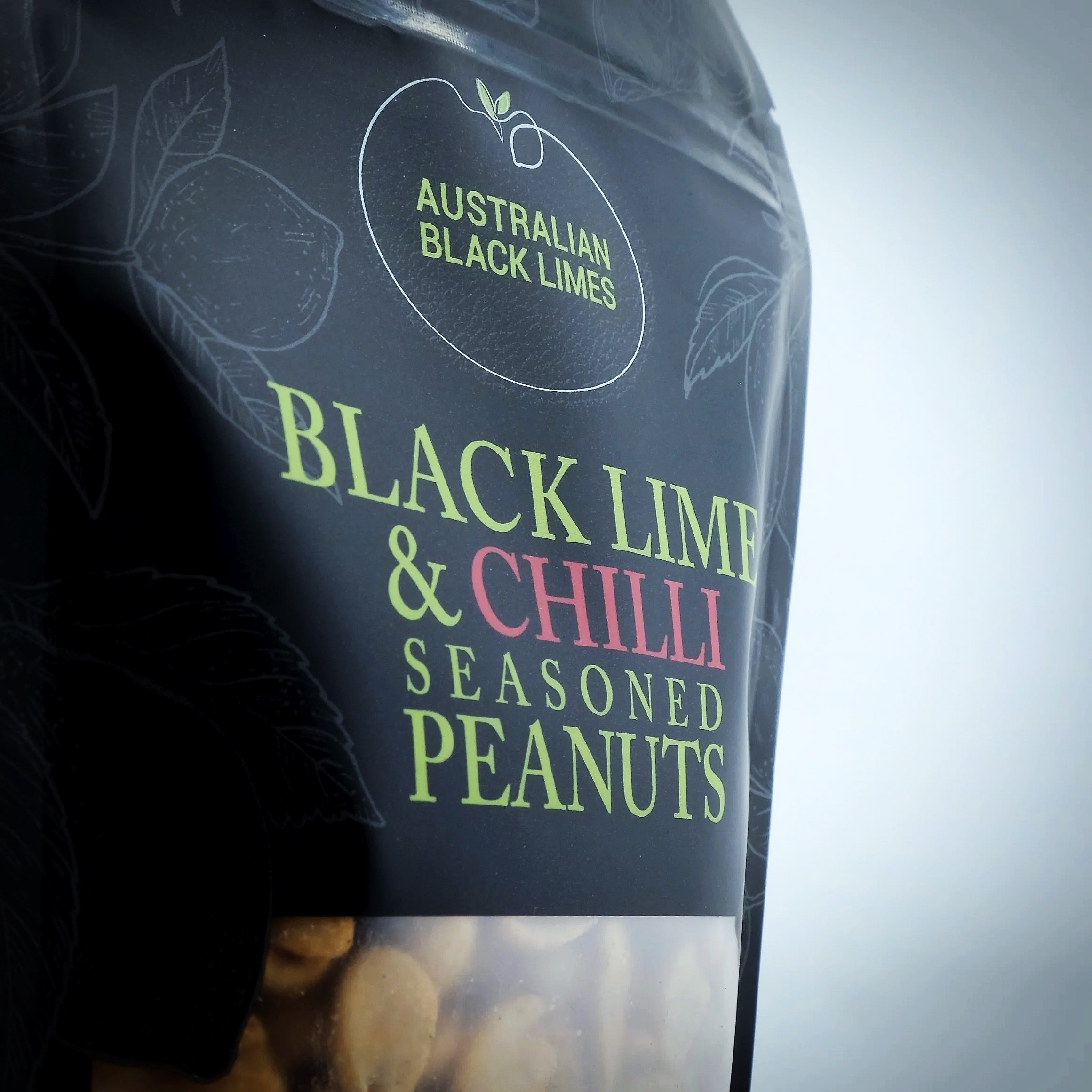 A close-up of a black surface, possibly a coffee cup or container, with colorful text that reads 'Australian Black Lime & Chilli Seasoned Peanuts' and a simple white outline of a lime to the top left of the text.