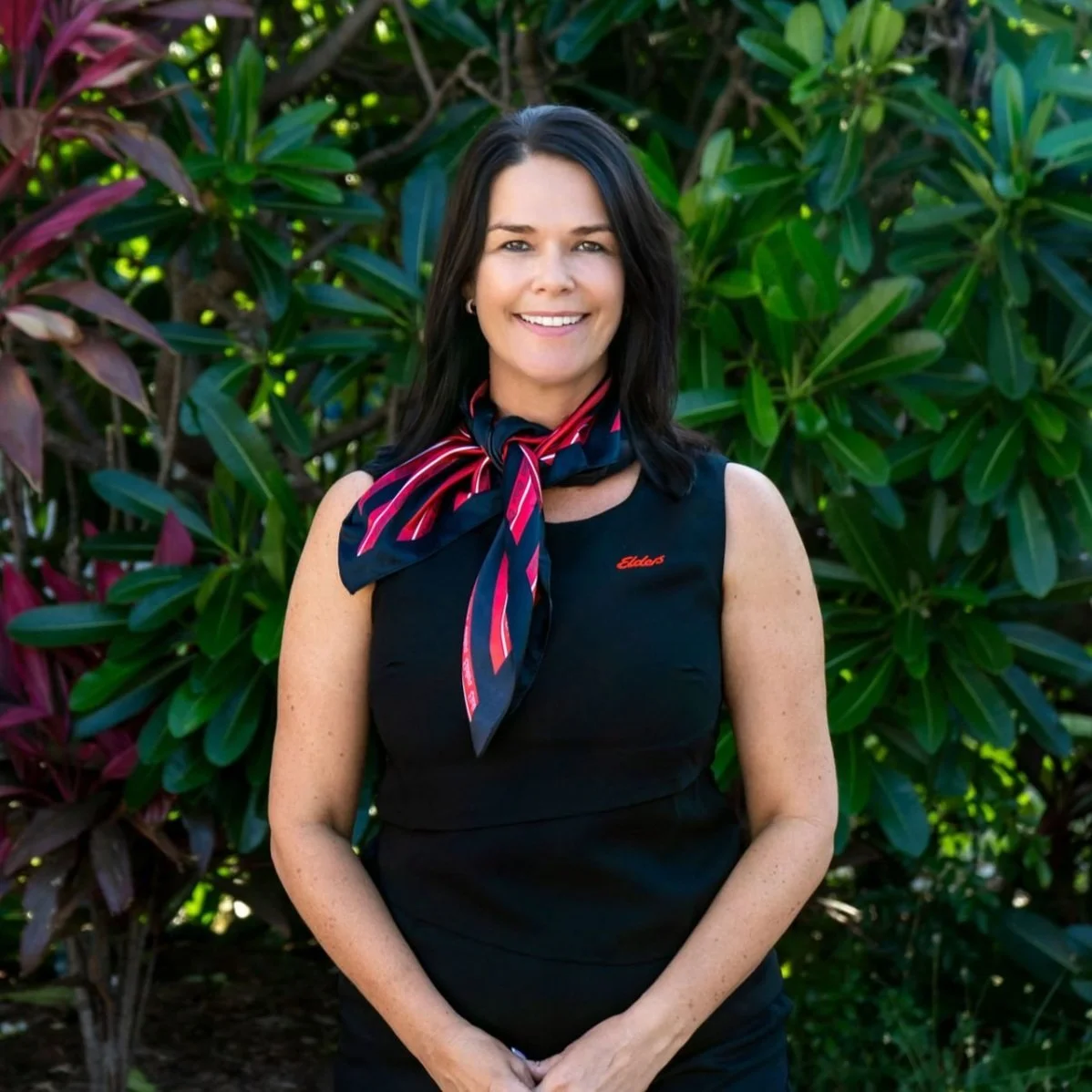 A woman with dark hair, wearing a sleeveless black top and a red and blue striped scarf around her neck, standing outdoors in front of green and purple foliage, smiling at the camera.