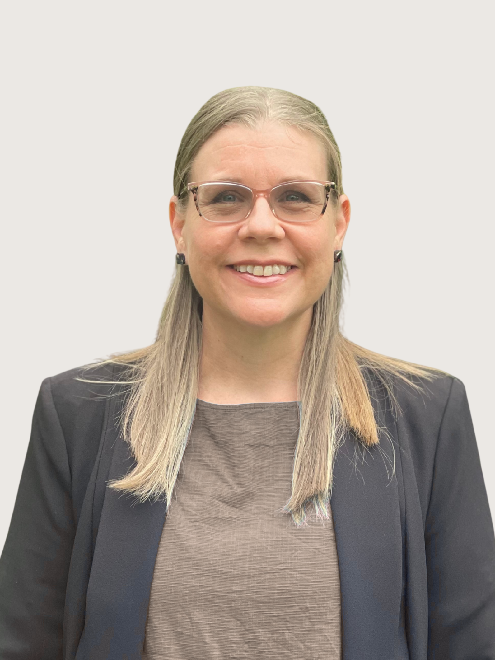 A woman with blonde hair wearing glasses, a dark blazer, and a light brown top, smiling against a light background.