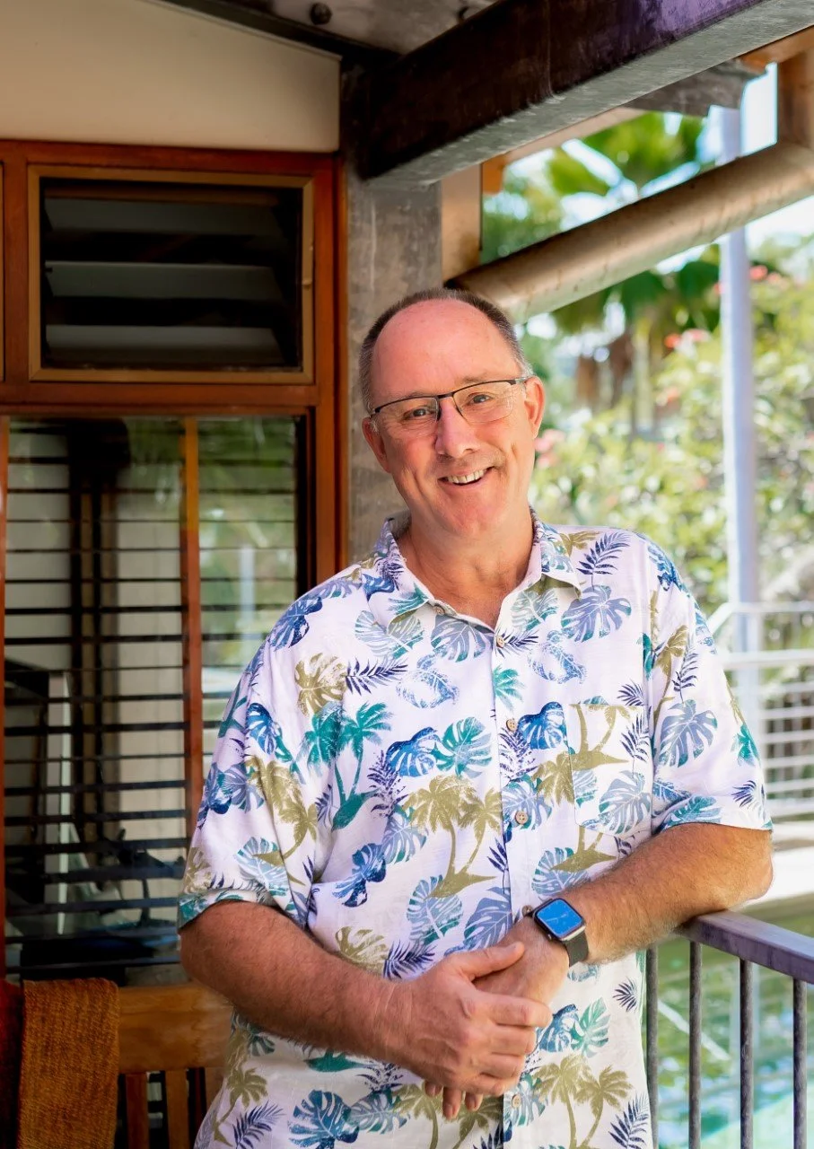 A smiling middle-aged man with glasses wearing a colorful tropical print short-sleeve shirt, standing on a balcony with greenery in the background.