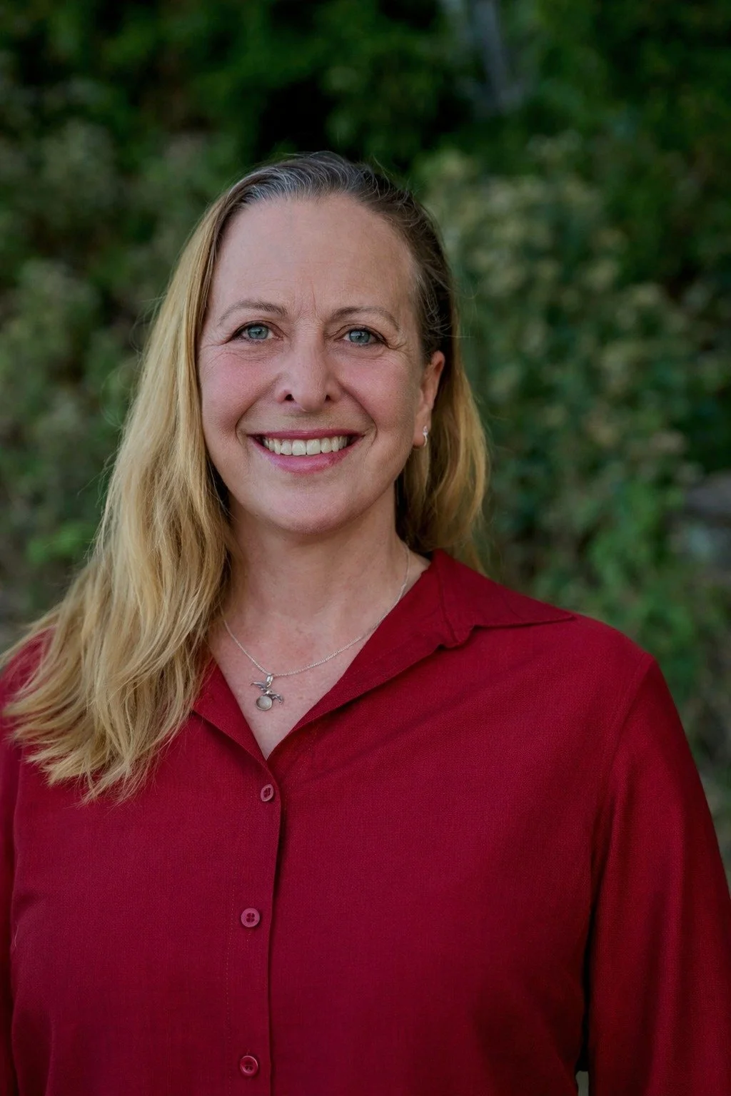 A smiling woman with blonde hair in loose waves, wearing a red button-up shirt and a silver necklace with a pendant, standing outdoors with a blurred green leafy background.