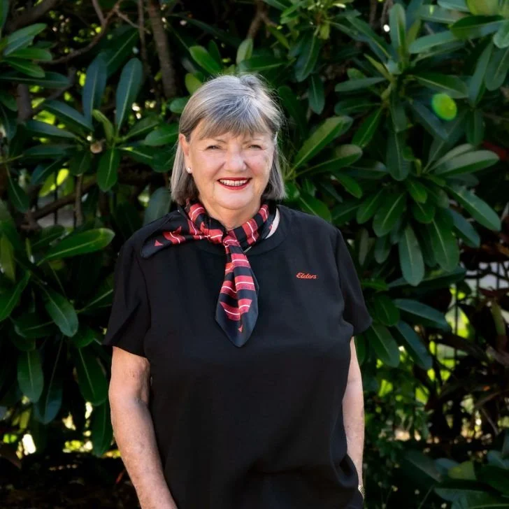 A smiling older woman with gray hair standing outdoors in front of green foliage, wearing a black shirt, a red and black patterned scarf, and a small red embroidered name tag that reads 'Eldon'.