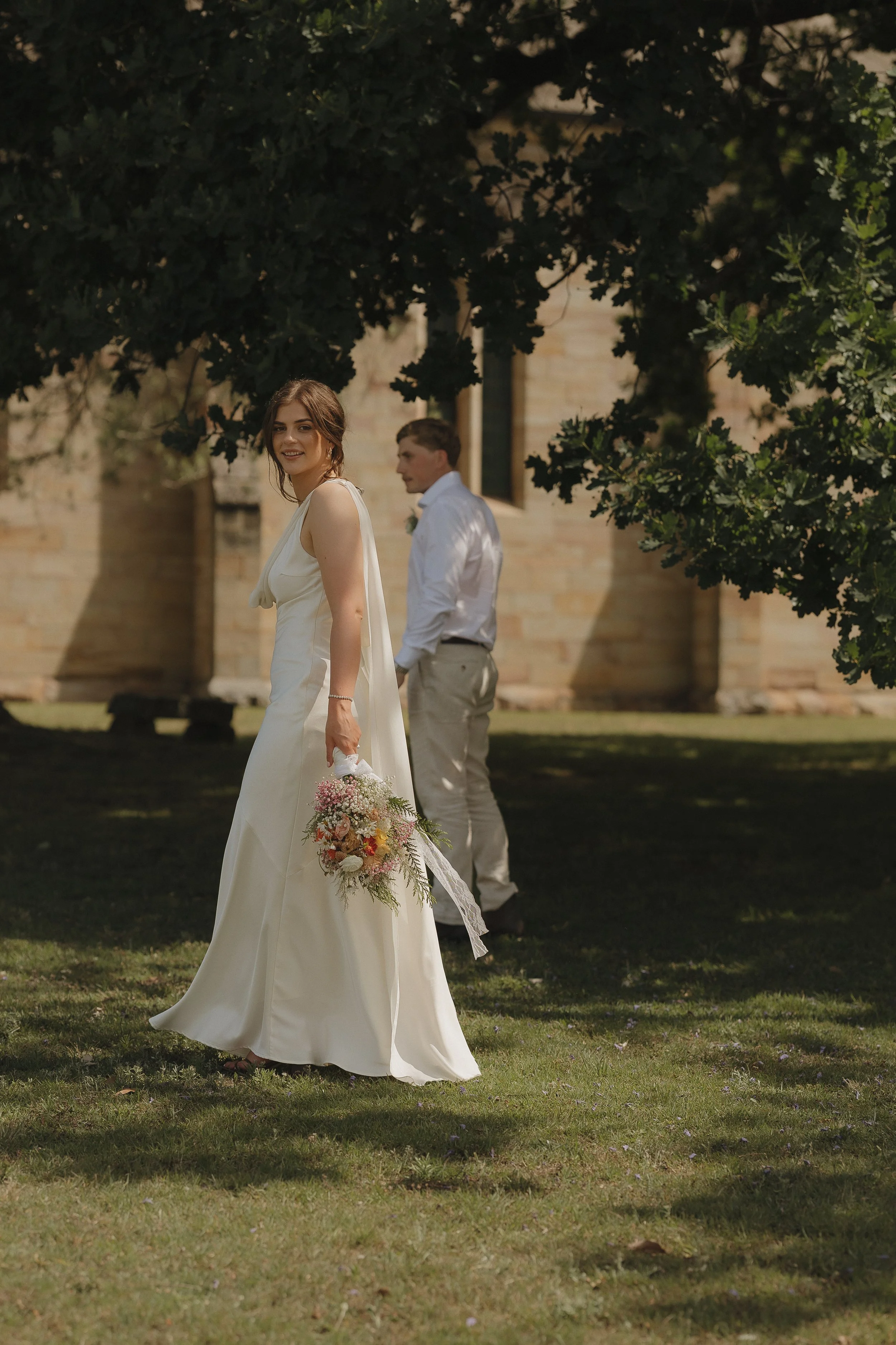 A bride in a white wedding dress holding a bouquet of flowers and smiling, standing outdoors under a tree; a groom in a white shirt and light-colored pants standing behind her, with a stone building in the background.