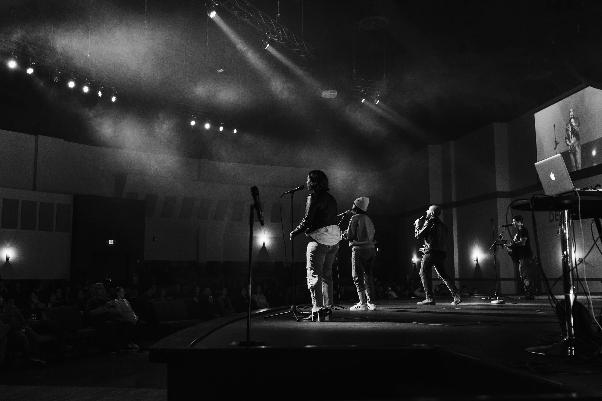 Black and white photo of a stage with five people singing into microphones, with the audience visible in the background.
