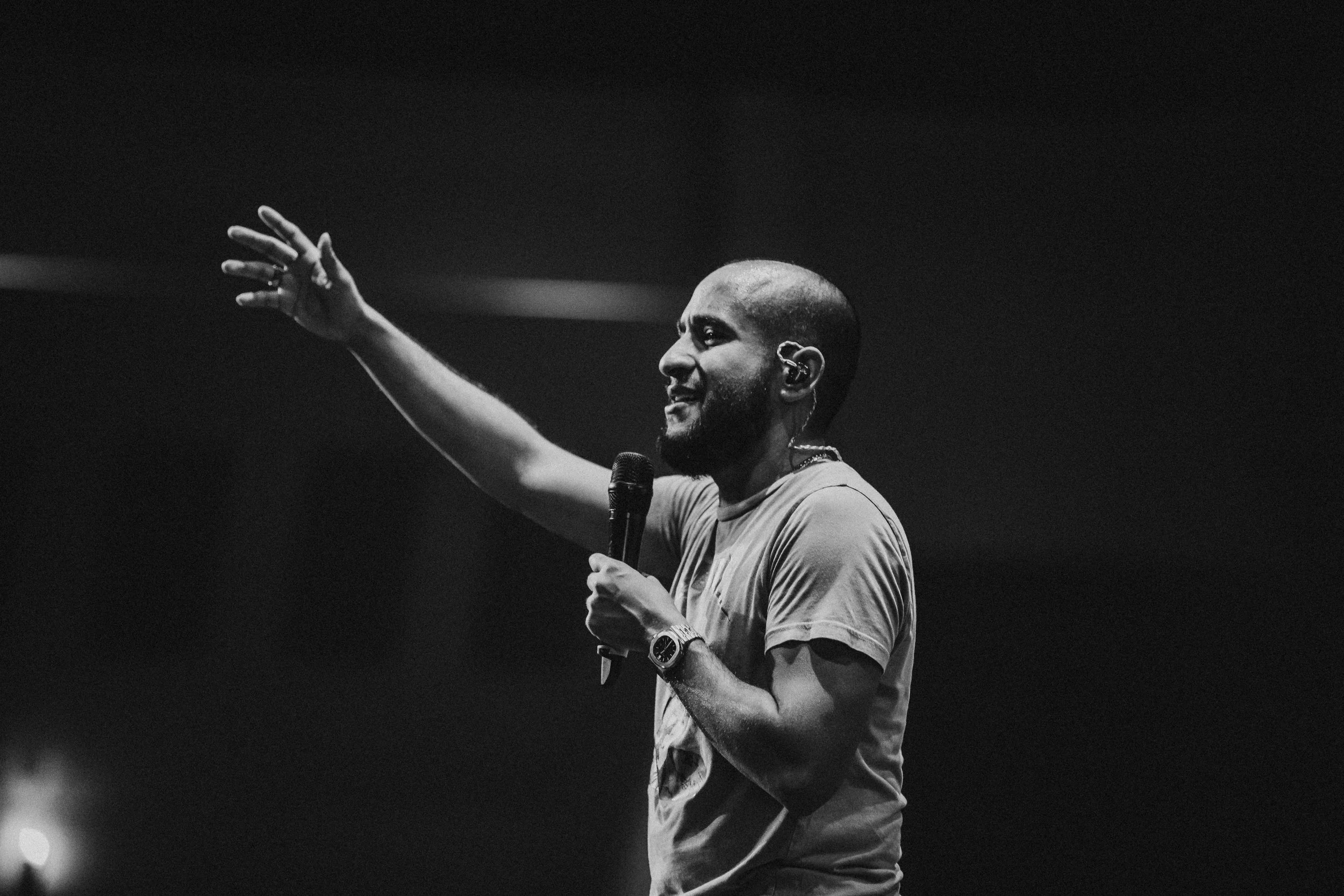 A man is speaking into a microphone on stage, gesturing with his right hand, in black and white.
