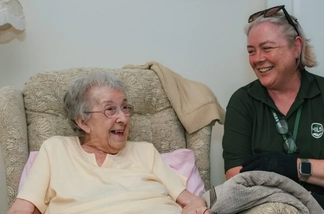 An elderly woman and a caregiver laughing together in a living room.