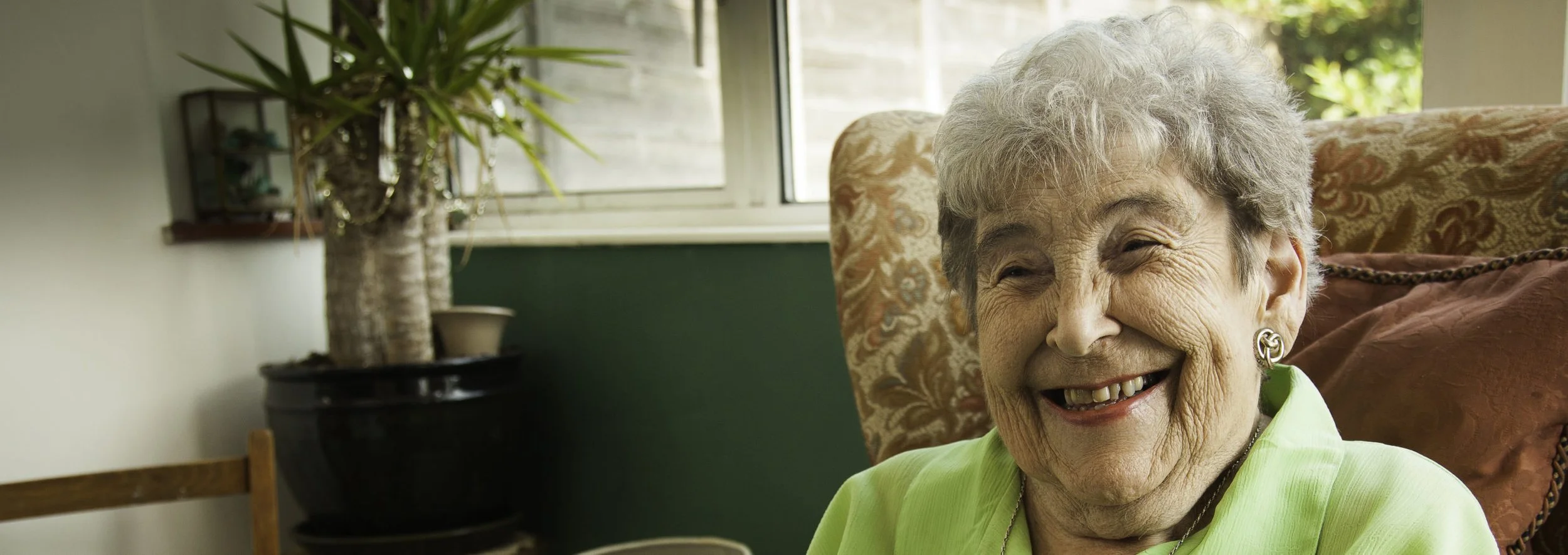 Smiling elderly woman with curly gray hair wearing a light green blouse, sitting in a cozy room with a patterned chair and a large potted plant near a window.