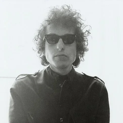 A black and white photo of the singer-songwriter and folk singer Bob Dylan from around 1965. He has curly hair, is wearing black Ray-Ban sunglasses and a dark shirt and is looking directly into the camera.