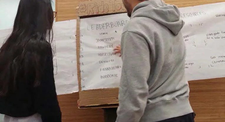 Two people looking at a large sheet of paper with handwritten notes taped to a wooden wall.