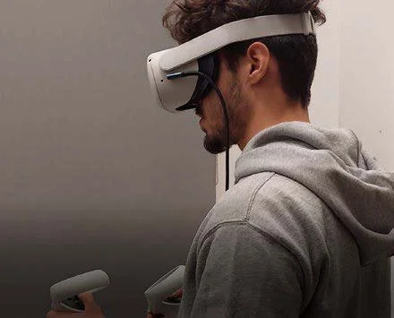 A man wearing a gray hoodie and a virtual reality headset using handheld controllers.
