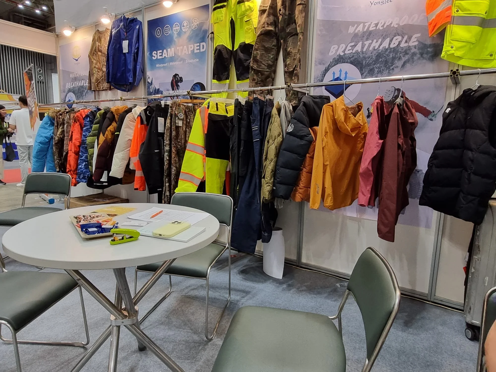 A trade show booth showcasing outdoor and weather-resistant clothing including jackets, pants, and vests in various colors and patterns. The booth features signage highlighting waterproof, windproof, and breathable features, with some garments hanging on a rack and posters on the wall. There are tables and chairs in the foreground with pamphlets, a phone, and other promotional materials.