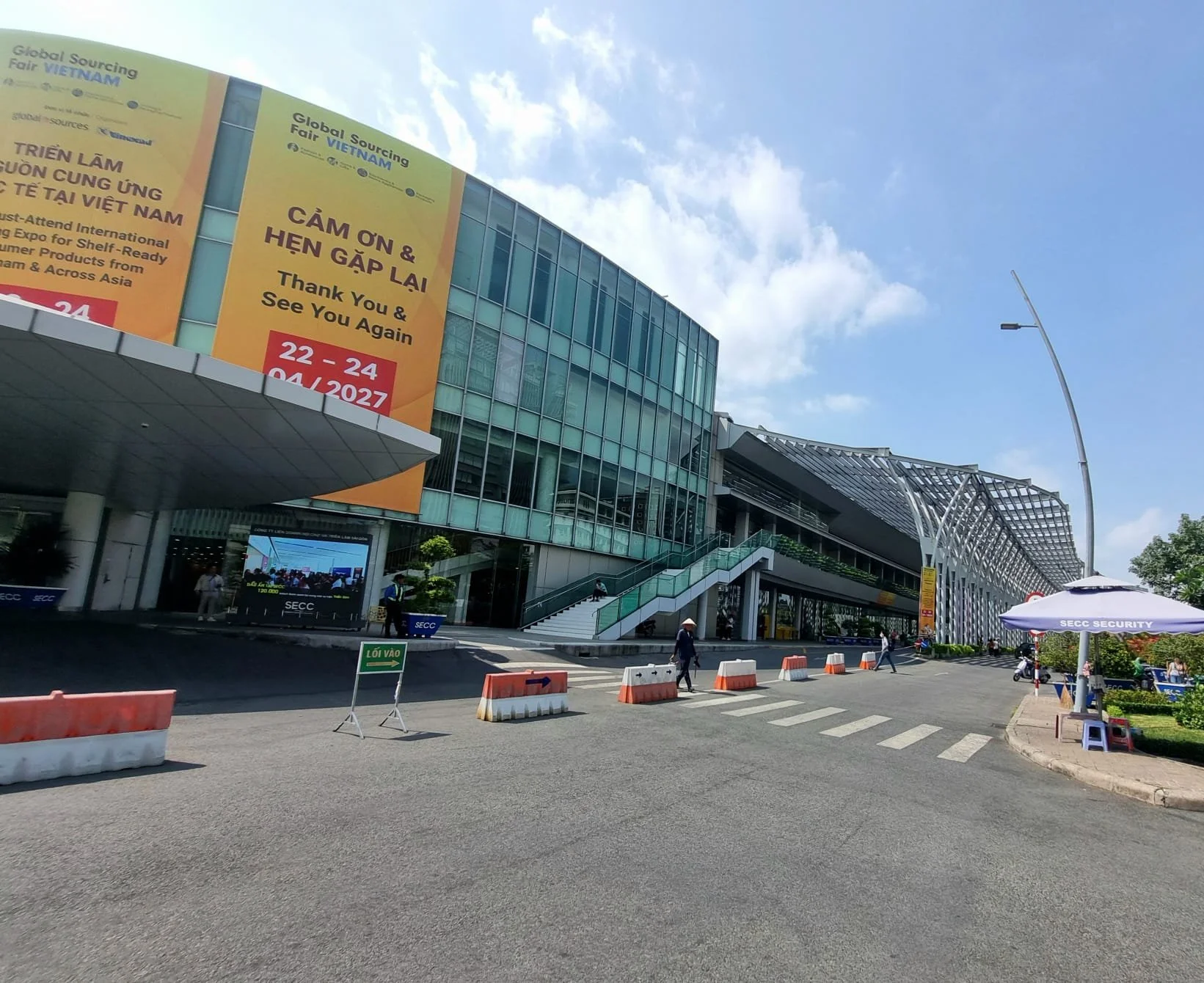 Exterior view of a modern building with large glass windows, a staircase, and yellow banners for the Vietnam Global Sourcing Fair, with people walking and the street scene including barriers and security.