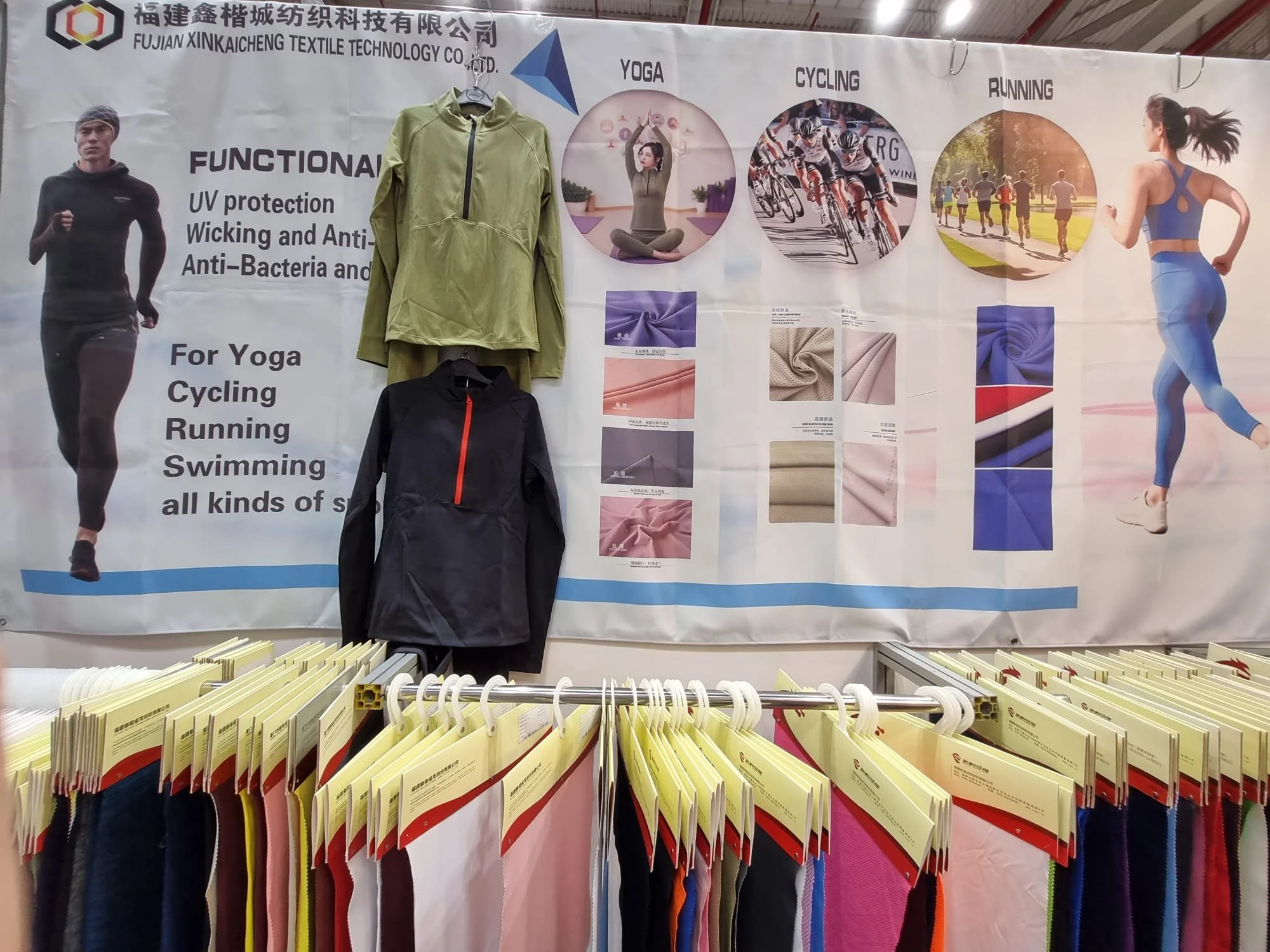 A large textile company's booth displays fabric and clothing samples with promotional poster in the background. The poster features images of people engaging in yoga, cycling, running, and swimming, highlighting the clothing's functions such as UV protection, antibacterial, and anti-wicking properties.