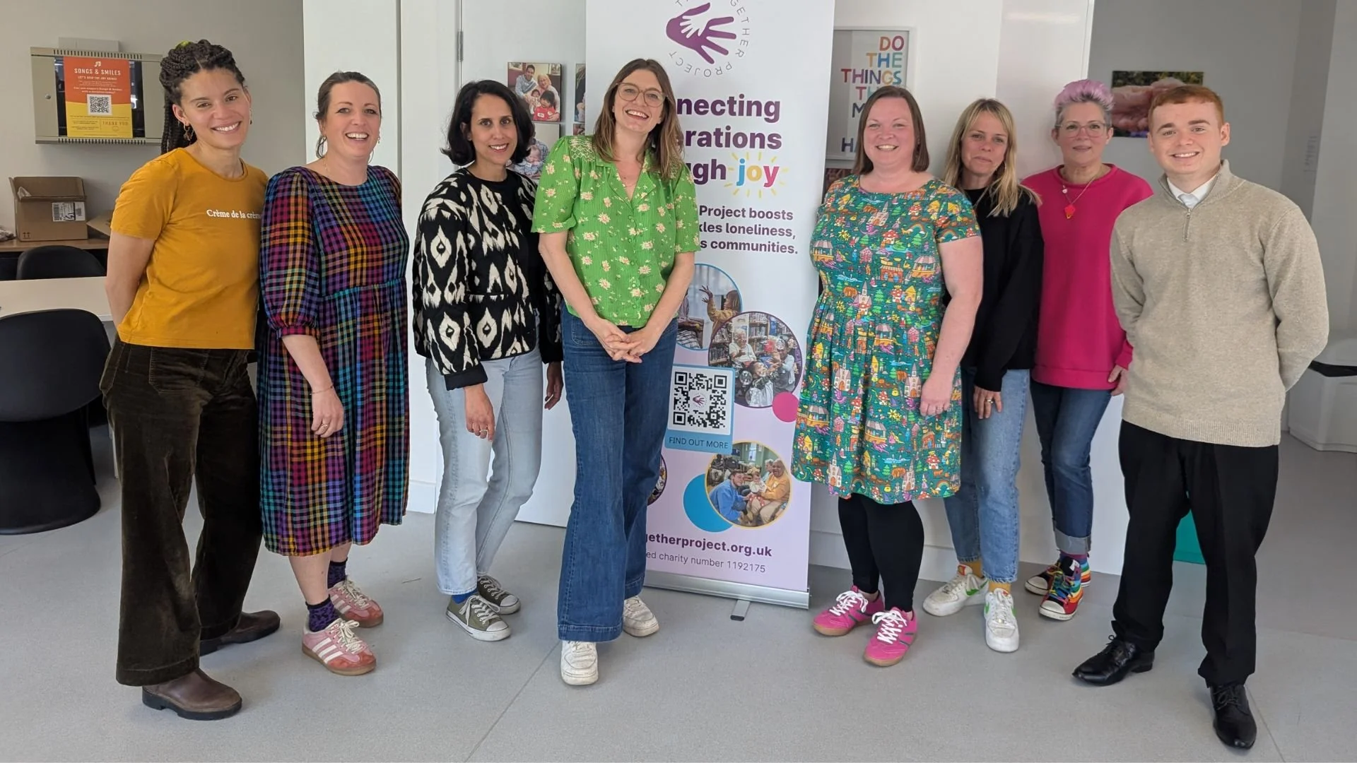 The Together Project team smiling together indoors beside a banner that reads ‘Connecting generations through joy’. The banner features the charity’s logo and photos from Songs & Smiles sessions.