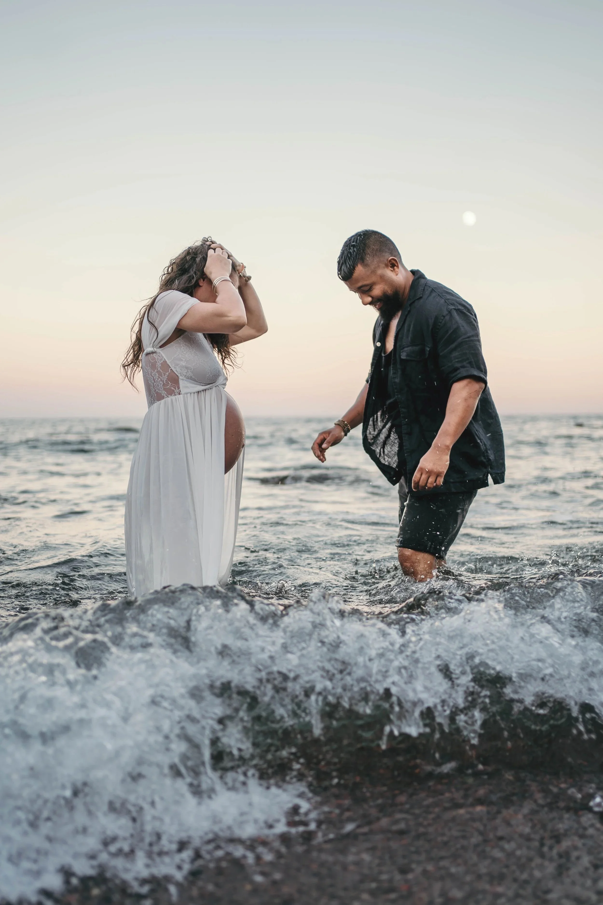 Un couple lors d'une séance photo sur la plage au coucher du soleil, la femme enceinte portant une robe blanche et l'homme en vêtements sombres, joyeux dans les vagues.