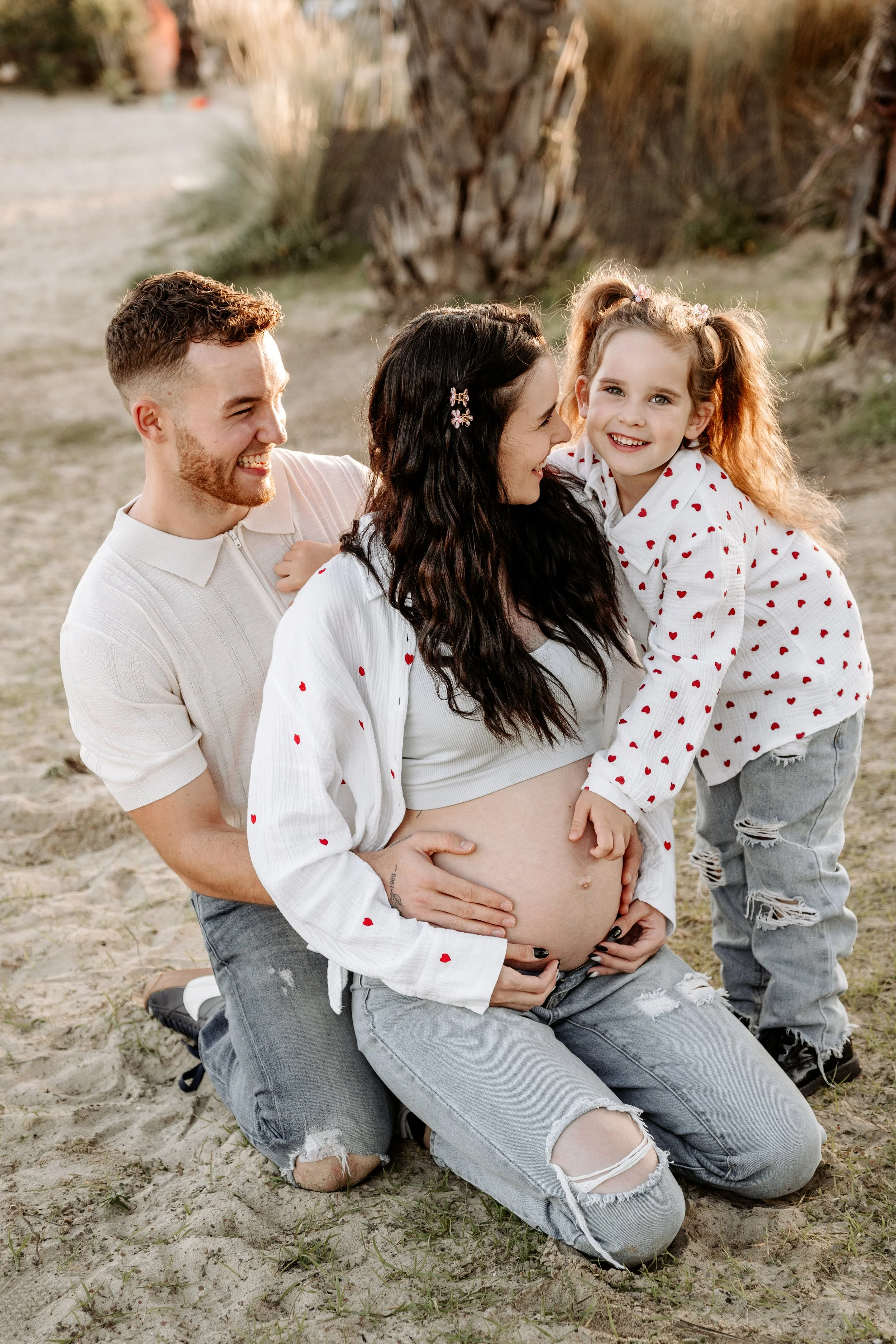 Une famille souriante composée d'un homme, d'une femme enceinte et d'une fille, posant sur la plage