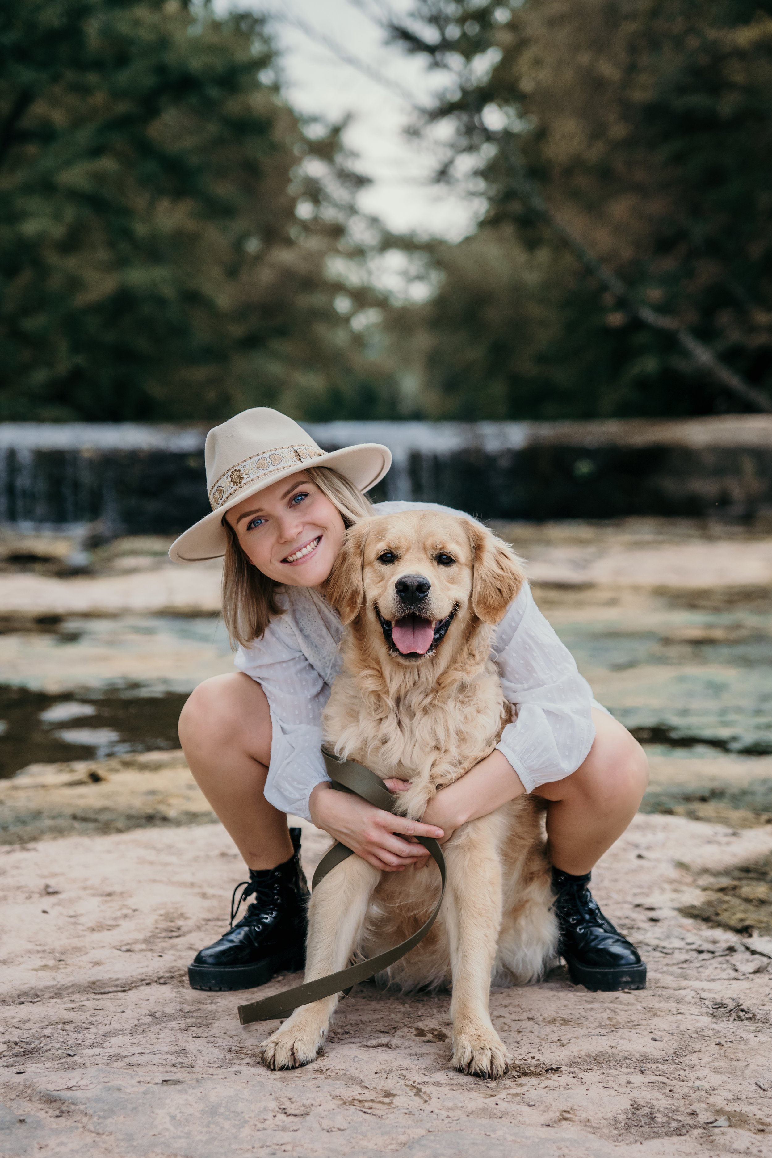 Une femme souriante portant un chapeau beige avec un chien Golden retriever jaune près d'une rivière dans la nature.