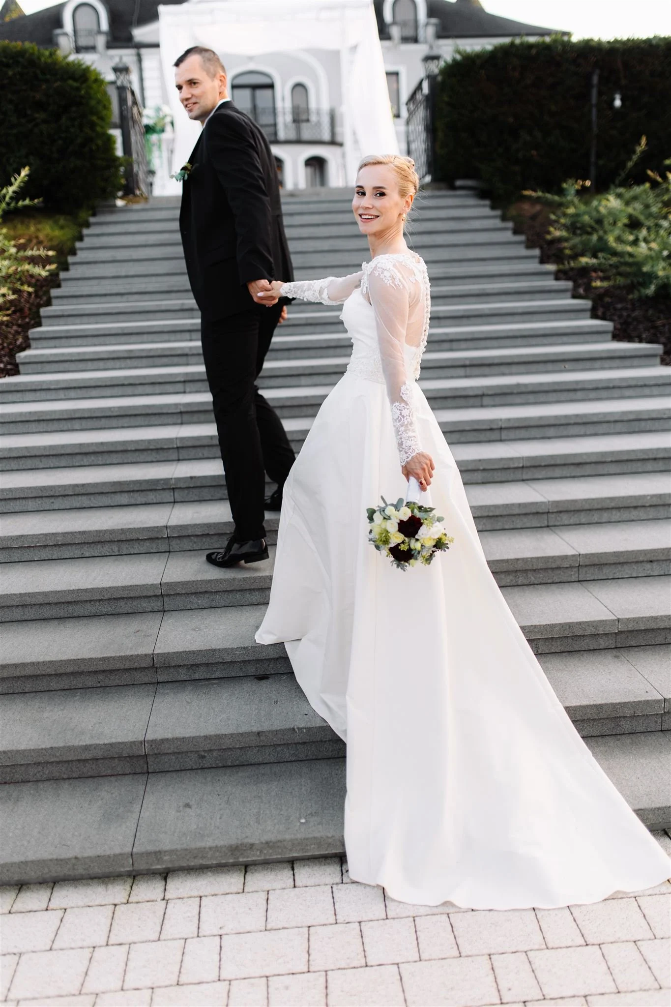 A bride and groom on outdoor steps in front of a white building; the bride, in a white wedding dress, holds a bouquet and looks back smiling, while the groom, in a black suit, looks at her.