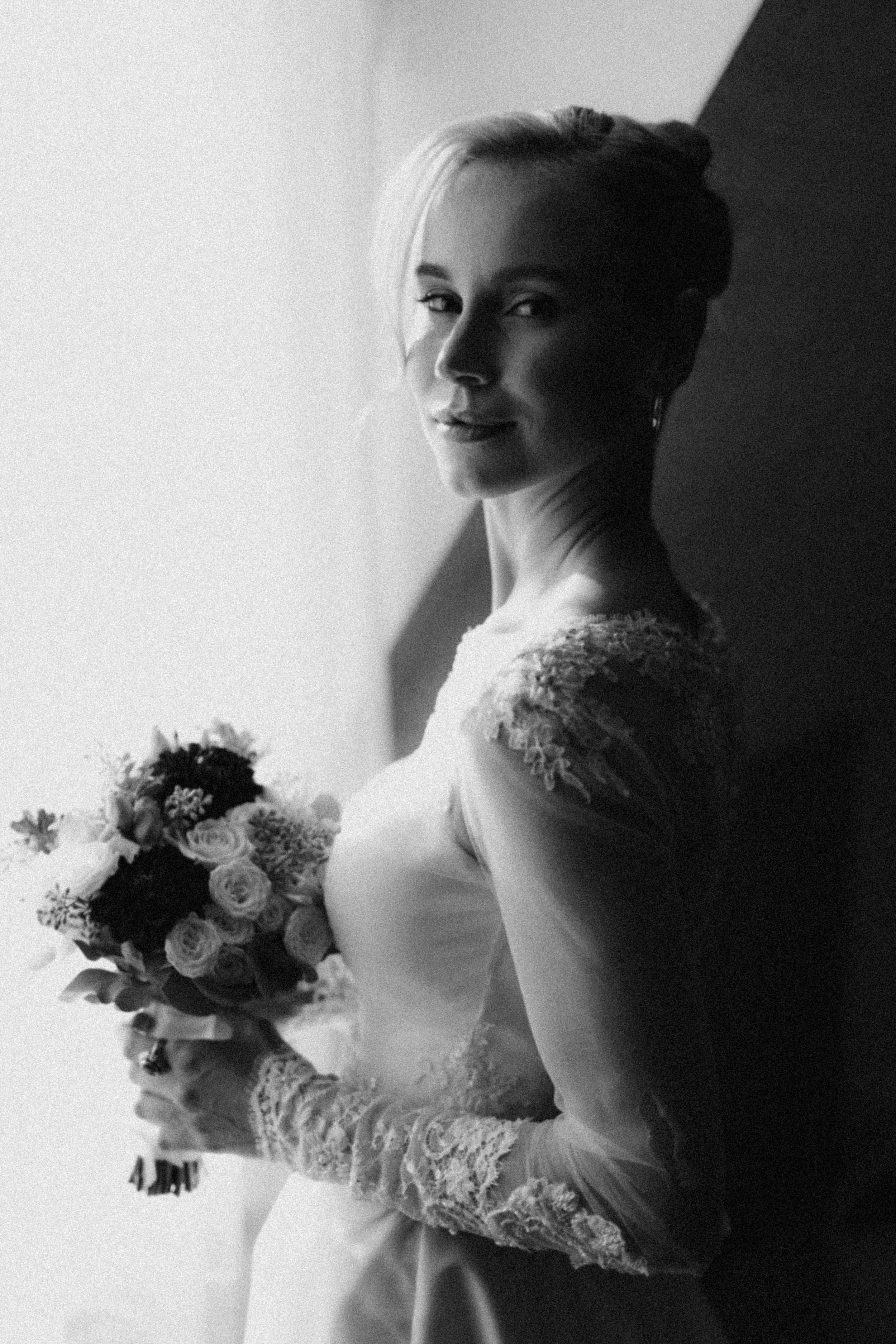 Black and white photo of a bride in a wedding dress with lace sleeves, holding a bouquet of flowers, standing near a window with sunlight.