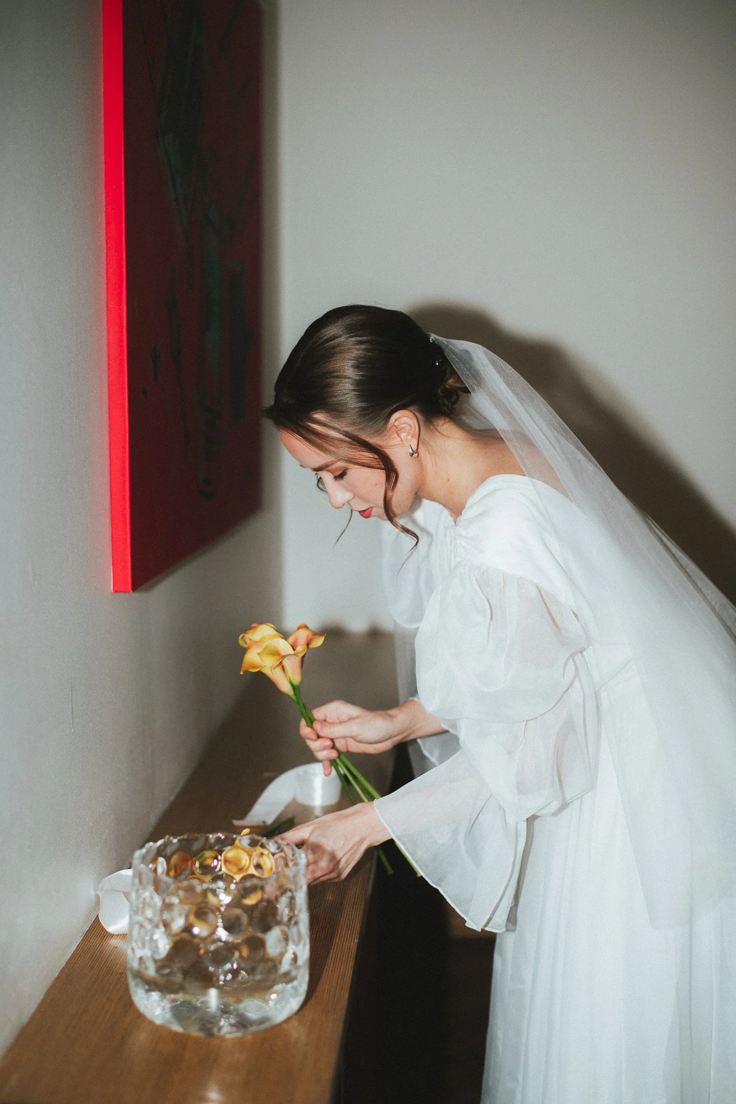 A bride in a white wedding dress and veil placing a yellow flower into a clear ice bowl on a wooden table.