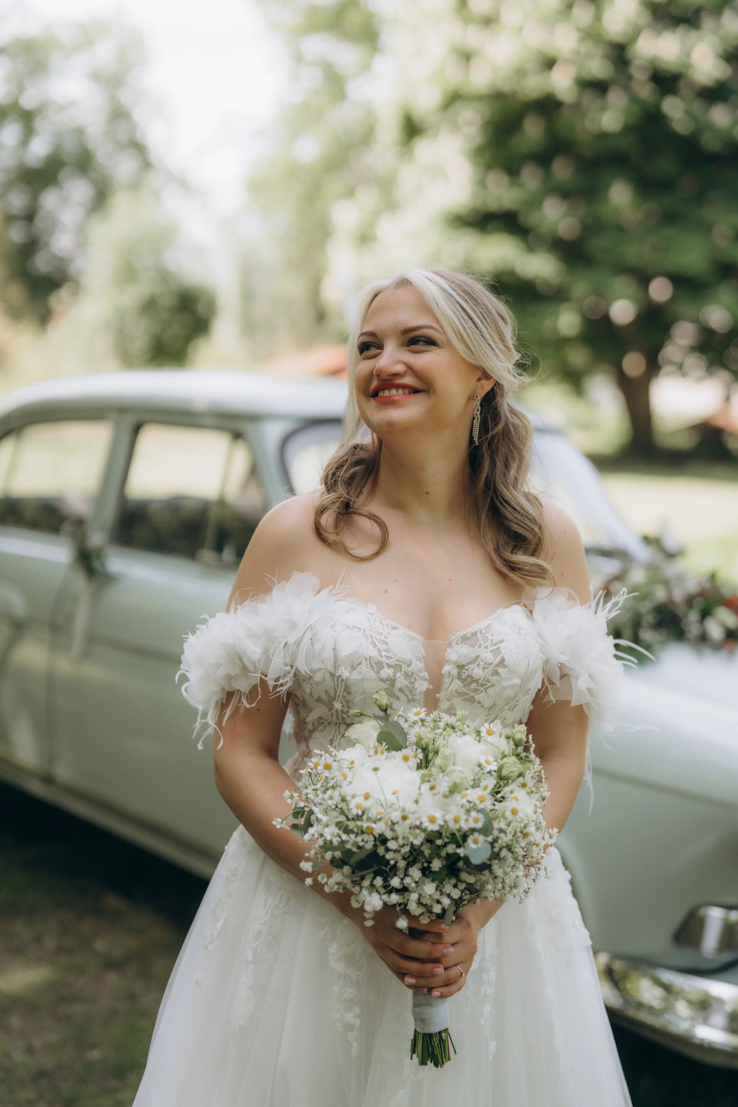 A bride in a white wedding gown holding a bouquet of white flowers, standing outdoors next to a vintage car with a background of trees and greenery.
