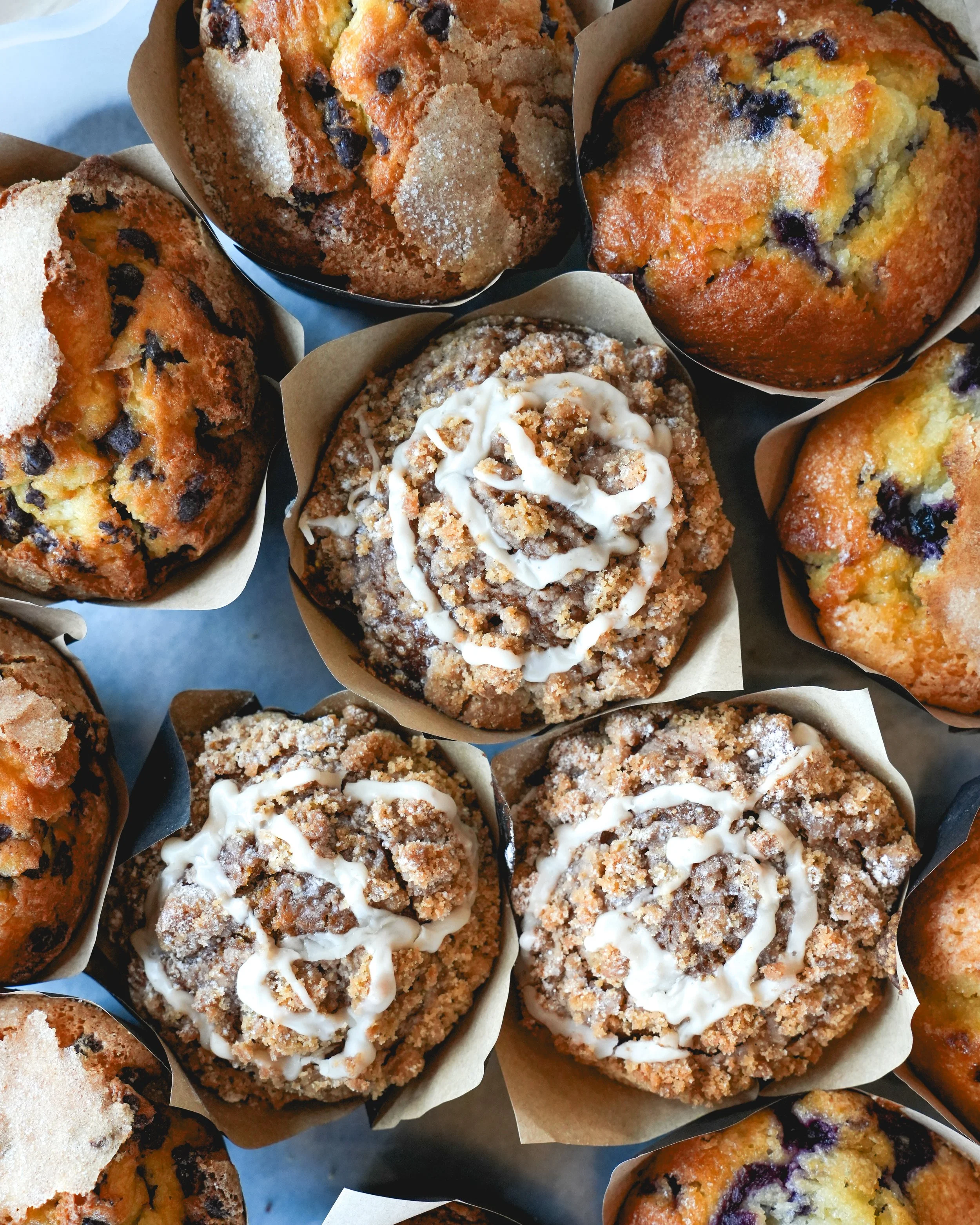 Close-up of assorted muffins, including blueberry muffins, with some topped with white icing and sugar, arranged on a tray.
