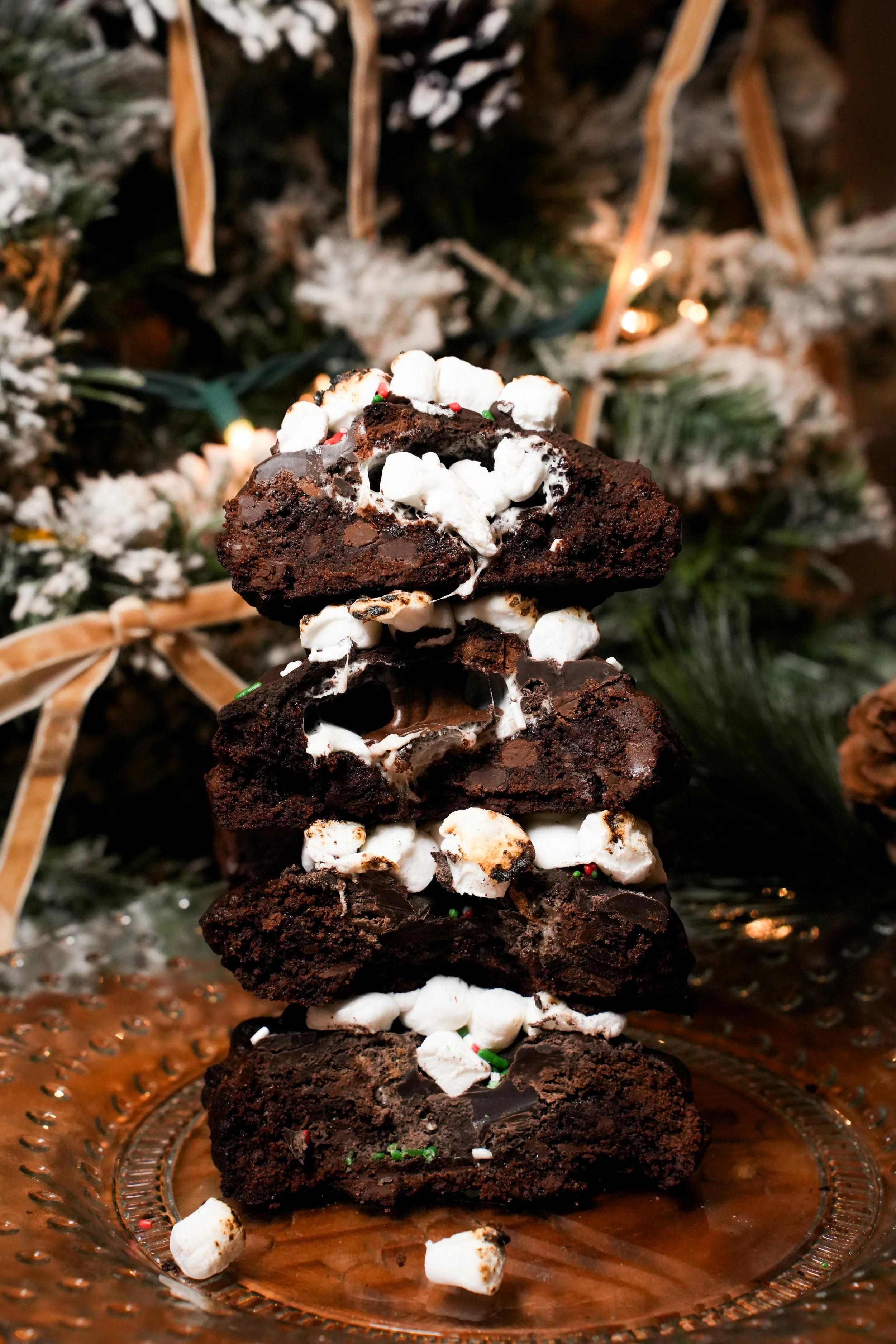 A stack of four chocolate brownies with marshmallows and sprinkles on top, set on a glass plate with a Christmas tree decorated with snow, pinecones, and ribbons in the background.