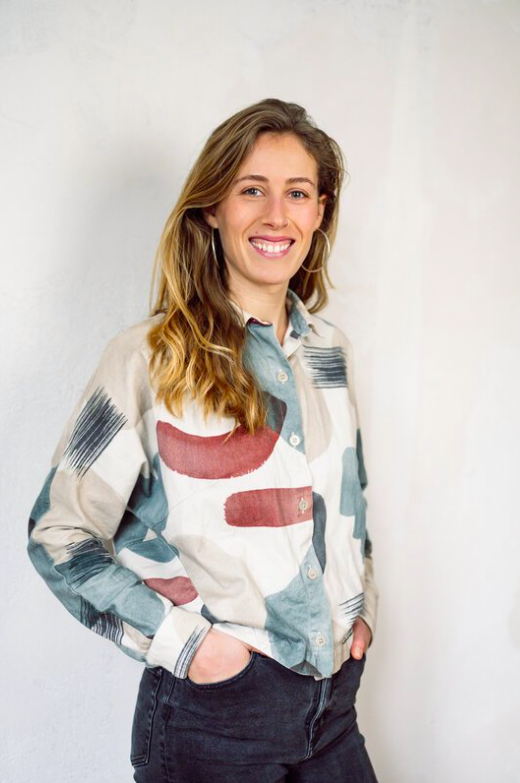 Junge Frau mit langem, gewelltem Haar, lächelnd, trägt eine bunt gemusterte Bluse und dunkle Jeans, steht vor weißer Wand.