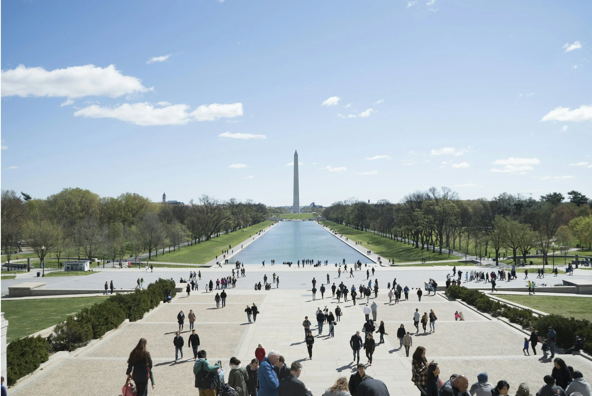 View of the National Mall in Washington, D.C., with the Washington Monument centered in the background, the Reflecting Pool stretching in front, and visitors walking along the steps and pathways.