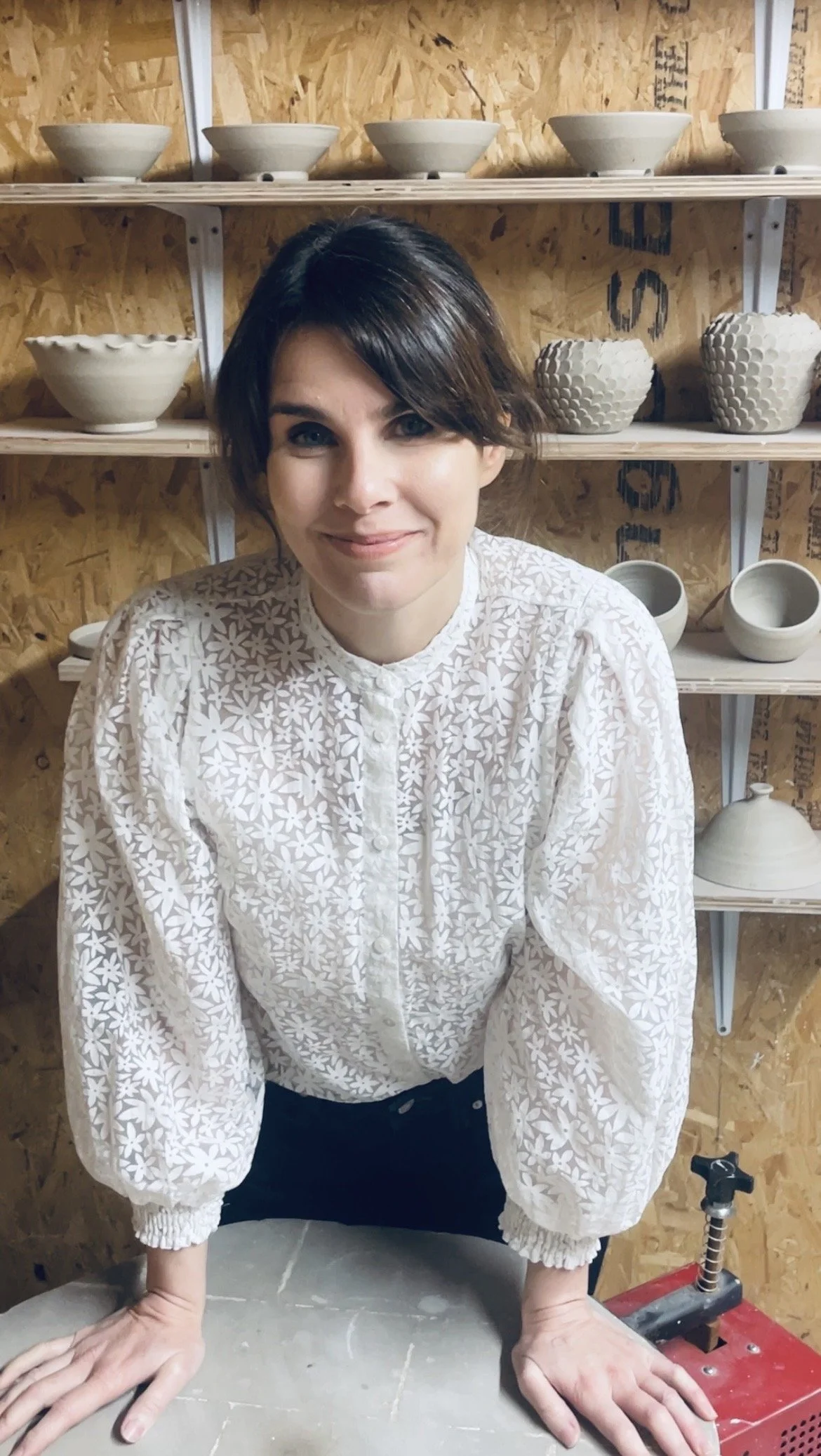 A woman with short dark hair smiling and leaning forward with hands on a table in a pottery studio. Shelves behind her hold various unglazed ceramic bowls and pots.