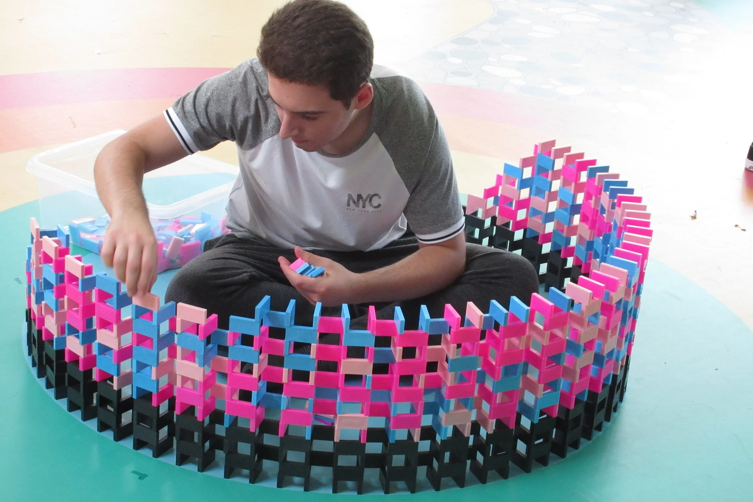 A young man sitting cross-legged on a teal floor, inside a circular enclosure made of black, pink, and blue dominoes. He is building this wall up around himself.