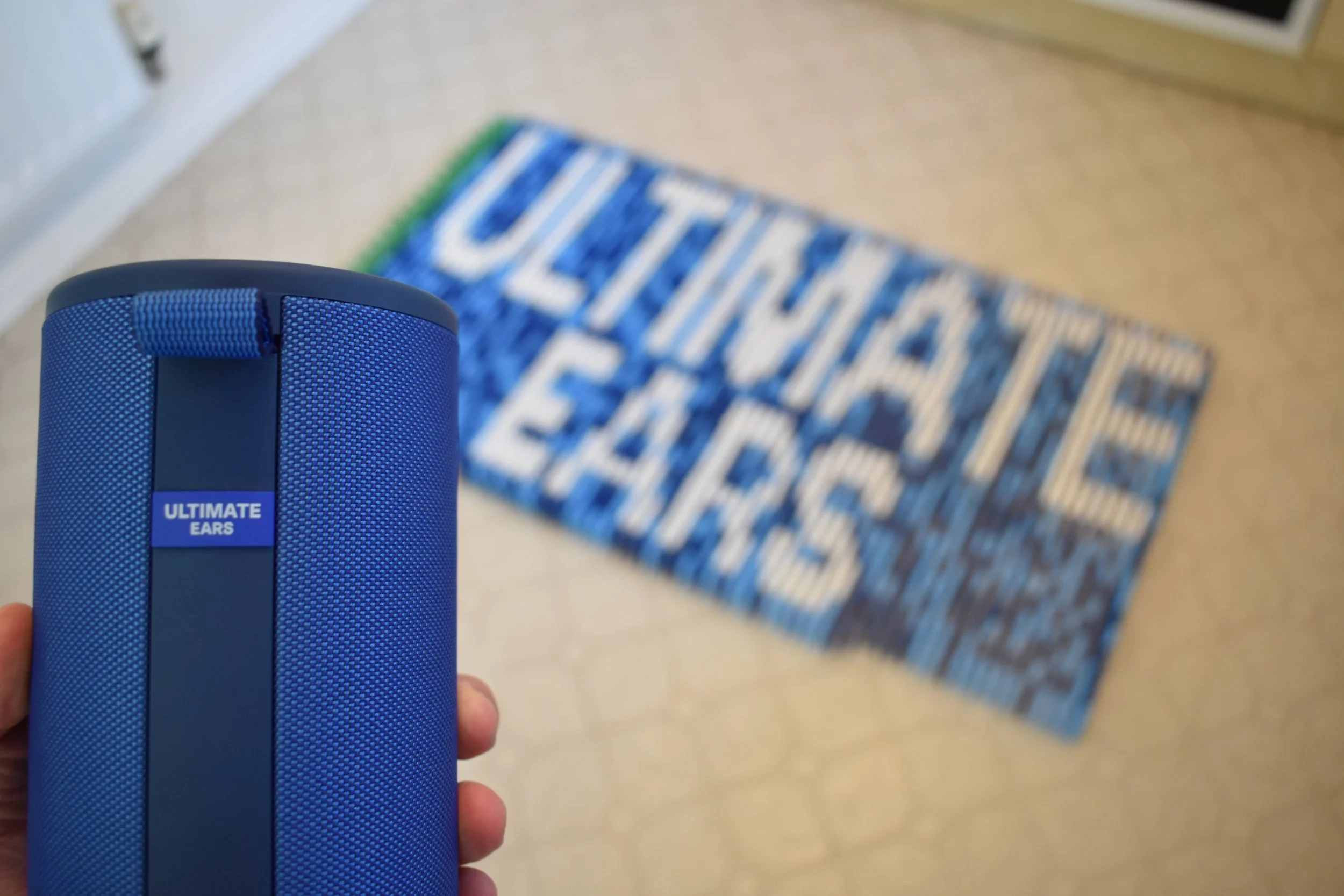 Close-up of a blue portable speaker labelled 'Ultimate Ears' held in a person's hand, with a blue and white mosaic in the background that says 'ULTIMATE EARS' in large letters.