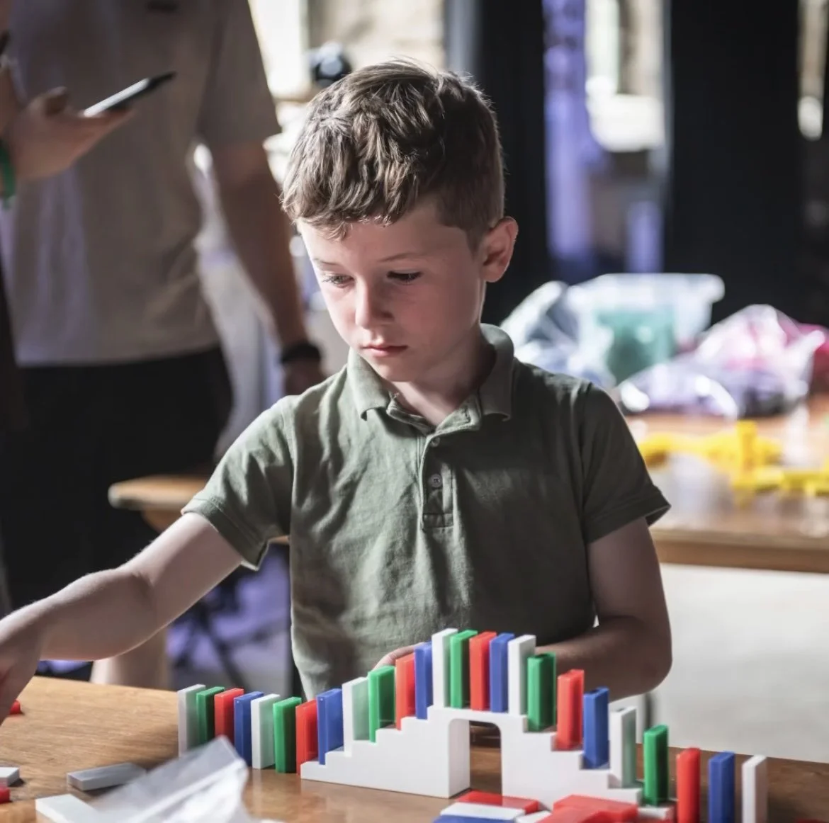 A young boy playing with colourful dominoes and a bridge on a wooden table indoors, with a few adults in the background.