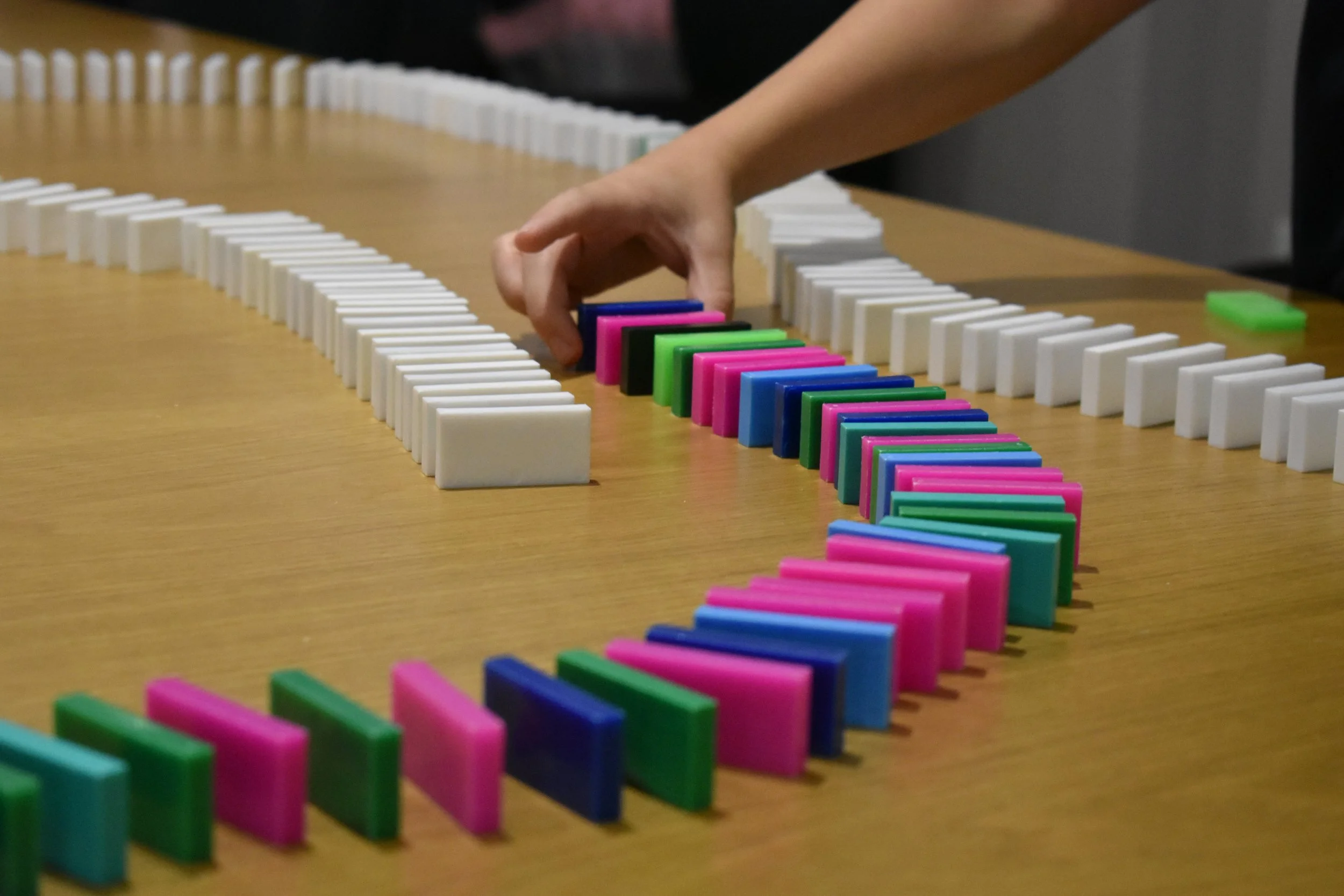 A person’s hand arranging dominoes on a wooden table, with dominoes set up horizontally. 